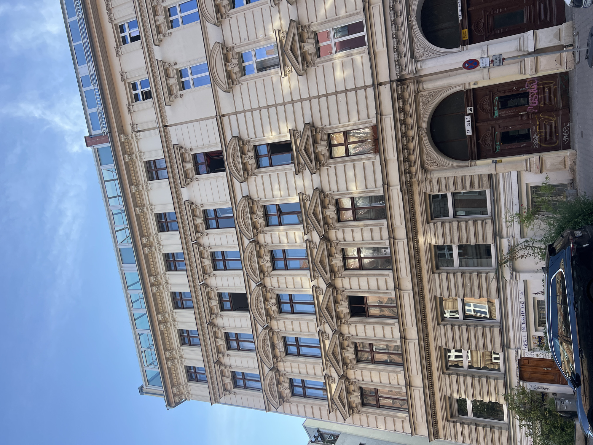 The image depicts the facade of a multi-story building in Berlin, Germany, under a clear blue sky with scattered clouds. The building exhibits classic European architectural elements, with ornate detailing around the windows and cornices. The facade is a light, neutral color, likely cream or beige, and features numerous windows with dark brown frames. Many of the windows are multi-paned. The building has a modern glass-enclosed addition at the top.
At the ground level, there are two large, arched wooden doors, characteristic of older Berlin buildings. The doors are dark brown and appear weathered, with some graffiti visible on them. Above the left door, a sign reads "SOUTERRAIN 38 E". The number "38 38 A-B 38" is also visible on the archway of the left door. To the right of the doors, a street sign with a red circle and a blue bar, along with other German text, is mounted on a pole.
A dark-colored station wagon is parked in front of the building, partially obscuring the ground floor. Greenery, including small trees and bushes, is visible on the left side of the building, and some climbing plants are growing on the facade. The overall impression is a street scene in a historic part of Berlin.