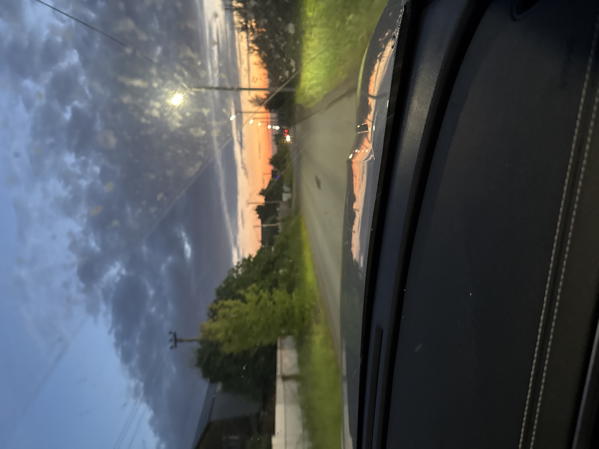 The image, taken from the interior of a vehicle, shows a road stretching into the distance under a twilight sky. The sky is a mix of deep blue and dramatic clouds, with a vibrant orange and yellow sunset visible on the horizon. Streetlights are intermittently visible along the road, some illuminated. On either side of the road, there is lush green grass and trees. In the background, a few houses and structures are visible, suggesting a semi-rural or suburban setting. The dashboard of the car is in the foreground, with its textured surface and stitching detail. The overall impression is one of a peaceful drive during dusk. There is no specific information or visual cues in the image to identify the location as Kazan, Russia.