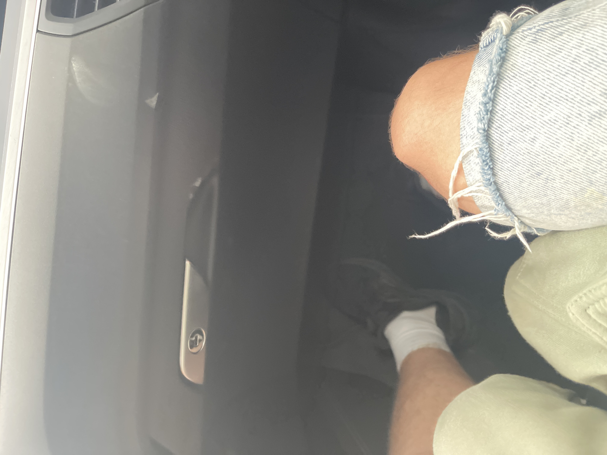 The image shows a person's legs and knees inside a vehicle. The person is wearing light green shorts and ripped denim jeans, with white socks and dark shoes. In the background, a car's dashboard is visible with a glove compartment latch. The scene appears to be an interior view of a car. There is no indication of the city of Kyiv, Ukraine, or any specific environmental details that would place it there.