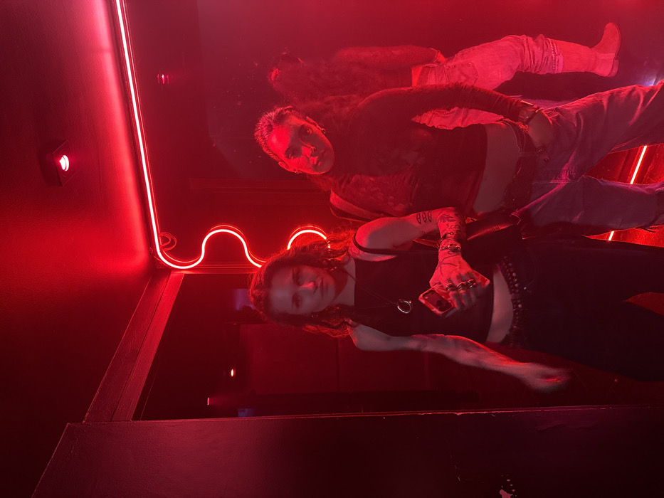 Two women in a dimly lit room under red lighting. One woman wears a black tank top, pants, and rings while holding a phone. The second woman is reflected above, wearing a long-sleeved top and jeans. A red neon light fixture is visible on the wall and ceiling. The scene appears to be in a bar or club setting.