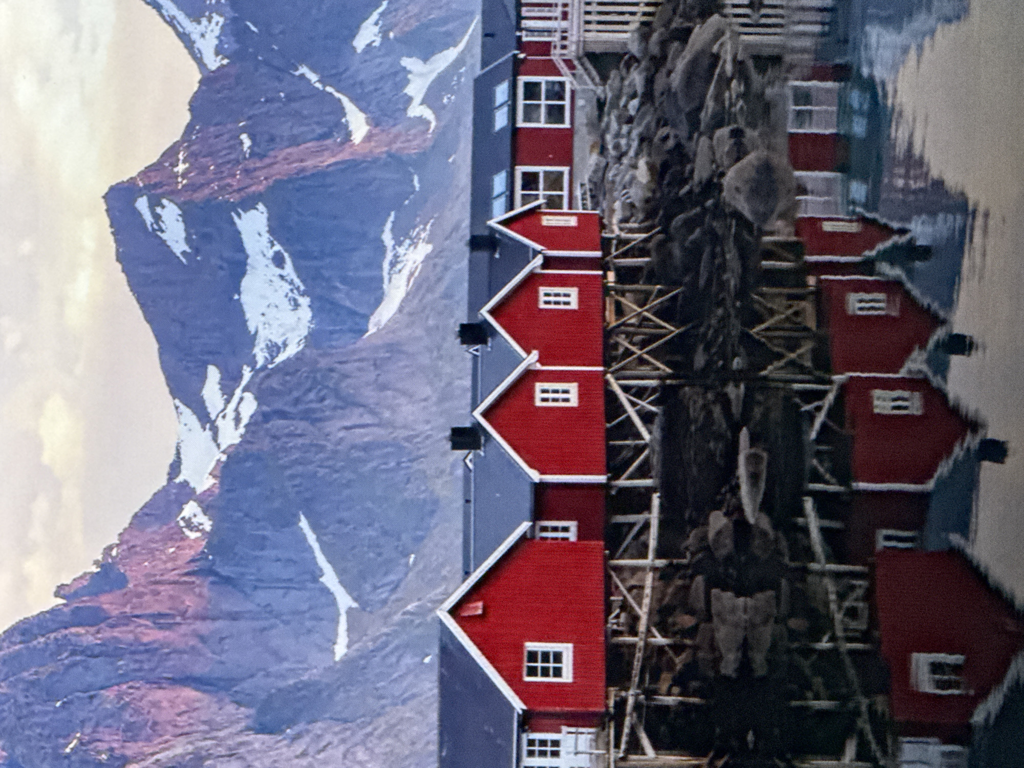 The image depicts a row of red buildings with white trim and blue roofs, reflected in the still water of a harbor or lake. The buildings are situated on a rocky shoreline, with a steep, snow-capped mountain range in the background. The mountain range is bathed in the warm, soft light of either sunrise or sunset, casting purple and pink hues on the snow and rock. The sky is a pale yellow, suggesting a calm and clear day. The water is perfectly still, creating a mirror image of the buildings and the surrounding landscape. There are no people visible in the image. The overall mood is serene and picturesque, evoking a sense of tranquility and natural beauty. The location appears to be a remote, coastal or lakeside community, possibly in a mountainous region like Norway or Alaska, given the architecture and natural landscape. There is no visible text in the image.