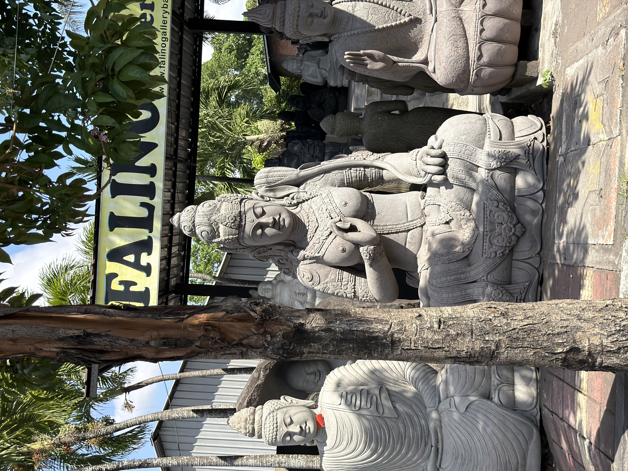 The image depicts an outdoor display of stone statues, likely for sale, in Denpasar, Indonesia. In the foreground, a large, intricately carved statue of a female deity, possibly an apsara or a Hindu goddess, is seated cross-legged with her hands in a devotional gesture. To the left of this central figure is a statue of a seated Buddha, also in a meditative pose. To the right of the central deity, another seated Buddha statue is visible, its right hand raised in a gesture of blessing or protection. Behind these primary statues, a variety of other stone sculptures, including smaller Buddha figures and possibly other deities, are arranged in the background. A tree trunk stands prominently in the middle of the frame, obscuring parts of the statues. Above the display, a yellow banner with the word "FALINO" in large black letters is visible, with "www.falinogallerybali.com" printed below. Palm trees and lush green foliage are seen behind the display, suggesting a tropical climate. The ground is paved with bricks and flagstones, and the scene is illuminated by bright sunlight, casting shadows from the statues and the tree. The overall impression is that of an outdoor gallery or showroom for stone carvings.