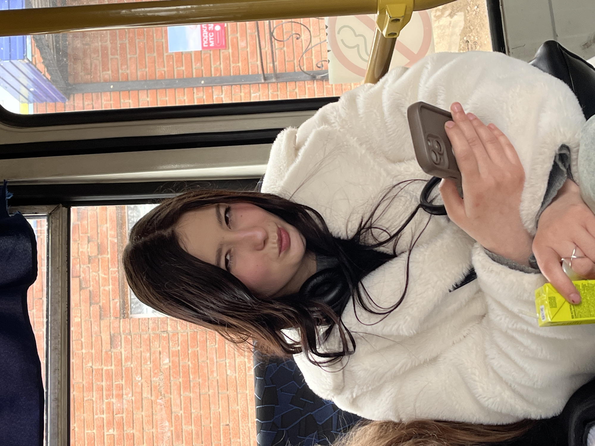 A young woman with long, dark hair is inside a bus. She is wearing a fluffy white jacket and headphones around her neck. She is holding a smartphone in her right hand and a small yellow juice box in her left. Outside the window, a brick building with signage in Russian is visible. The bus has a metal frame and patterned seats. Based on the signage, the location is identified as Фоки, Russia.