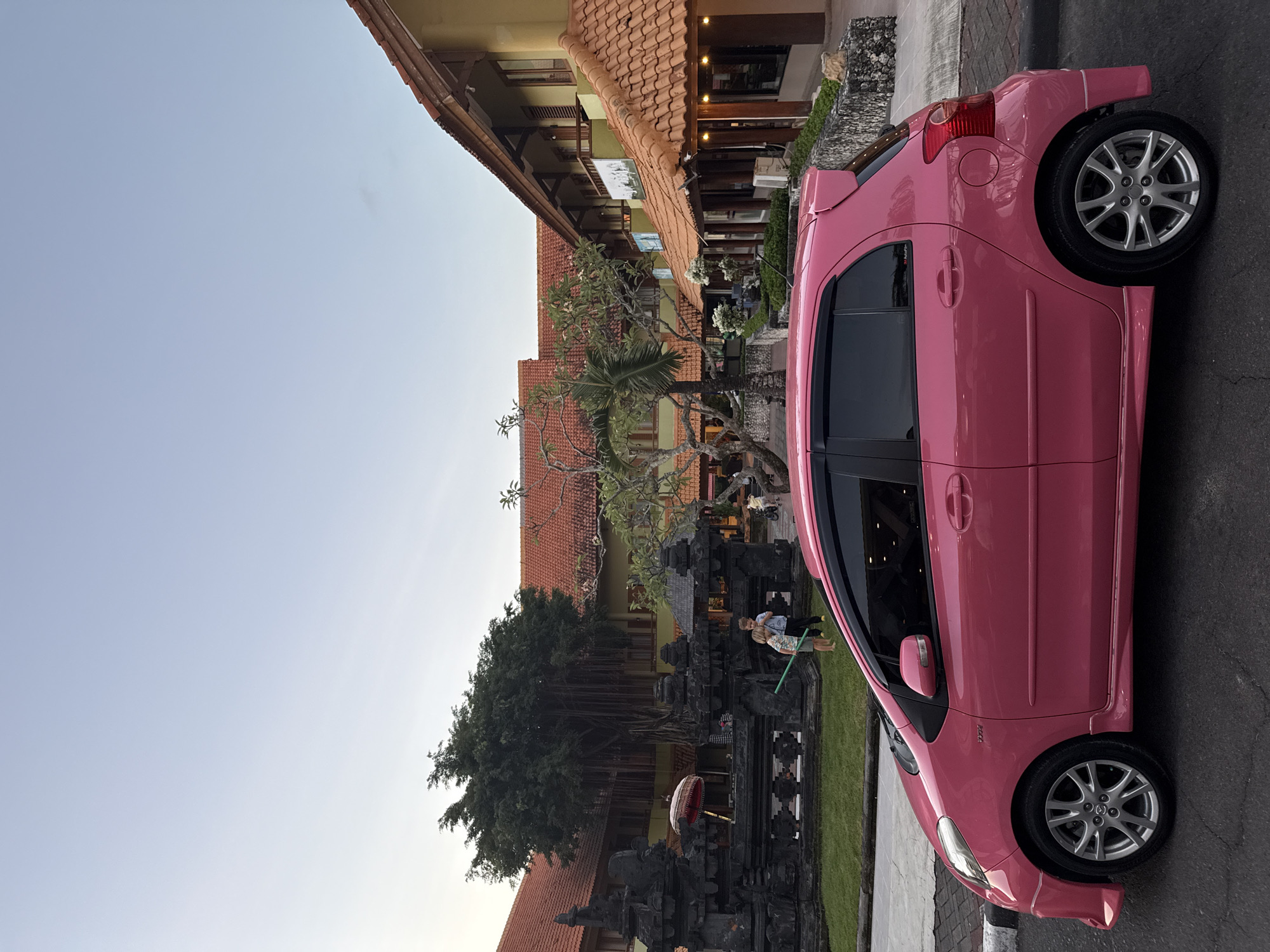 The image presents an outdoor scene depicting a vibrant pink hatchback car parked on a paved surface, likely a road or driveway, on the right side of the frame. The car, possibly a Toyota Yaris or similar model, faces away from the viewer and features multi-spoke silver alloy wheels.
To the left of the car, a lush tropical landscape unfolds, showcasing several buildings with traditional Balinese architecture. These structures are characterized by prominent orange-tiled roofs with multiple tiers and exposed wooden beams. Interspersed among the buildings and green lawns are various trees, including a large banyan-like tree with visible aerial roots, palm trees, and flowering plants, possibly frangipani.
In the mid-ground, dark, intricately carved stone structures, typical of Balinese temples or garden decorations, are visible. Near these structures, a traditional Balinese offering umbrella (payingung) with red and white patterns stands. Two individuals, possibly a child and an adult, are standing among the greenery and stone elements, with one person holding a long green stick or object.
The sky above is clear and a soft light blue, suggesting either late afternoon or early morning. Some artificial lights are visible within the buildings, hinting at the time of day being closer to dusk. The location cues, such as the architectural style, vegetation, and traditional artifacts, strongly indicate a setting in Bali, Indonesia, aligning with the provided context of Kuta Selatan. No legible text is visible in the image.