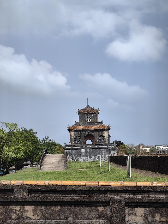 An exterior, eye-level photograph depicts the Hiển Lâm Các (Pavilion of Splendid Caprice) within the Imperial City, Phú Xuân Ward, Huế, Vietnam, under a partly cloudy sky. The central subject is a two-tiered, weathered stone and brick pavilion, characterized by its traditional tiered roof with reddish-brown tiles and dark grey, discolored walls. The lower tier features a central arched opening flanked by two circular, patterned windows. The upper tier displays a single circular, patterned window centrally placed. A small, indistinct human figure is partially visible on the lower tier to the left of the central archway.
In the immediate foreground, a dark grey stone wall with an orange-tiled coping runs horizontally across the bottom of the frame. Behind this wall, a strip of green grass precedes a concrete pathway ascending from the left towards the pavilion. To the right of the grass, a dirt path runs alongside the pavilion, marked by two light-colored vertical posts. Dense green trees and foliage are visible to the left of the pavilion. To the right, a dark stone wall or fortification extends into the midground, beyond which distant urban buildings with light-colored facades and various roof styles are discernible against the horizon. The sky is a light blue, containing scattered white and grey cumulus clouds.