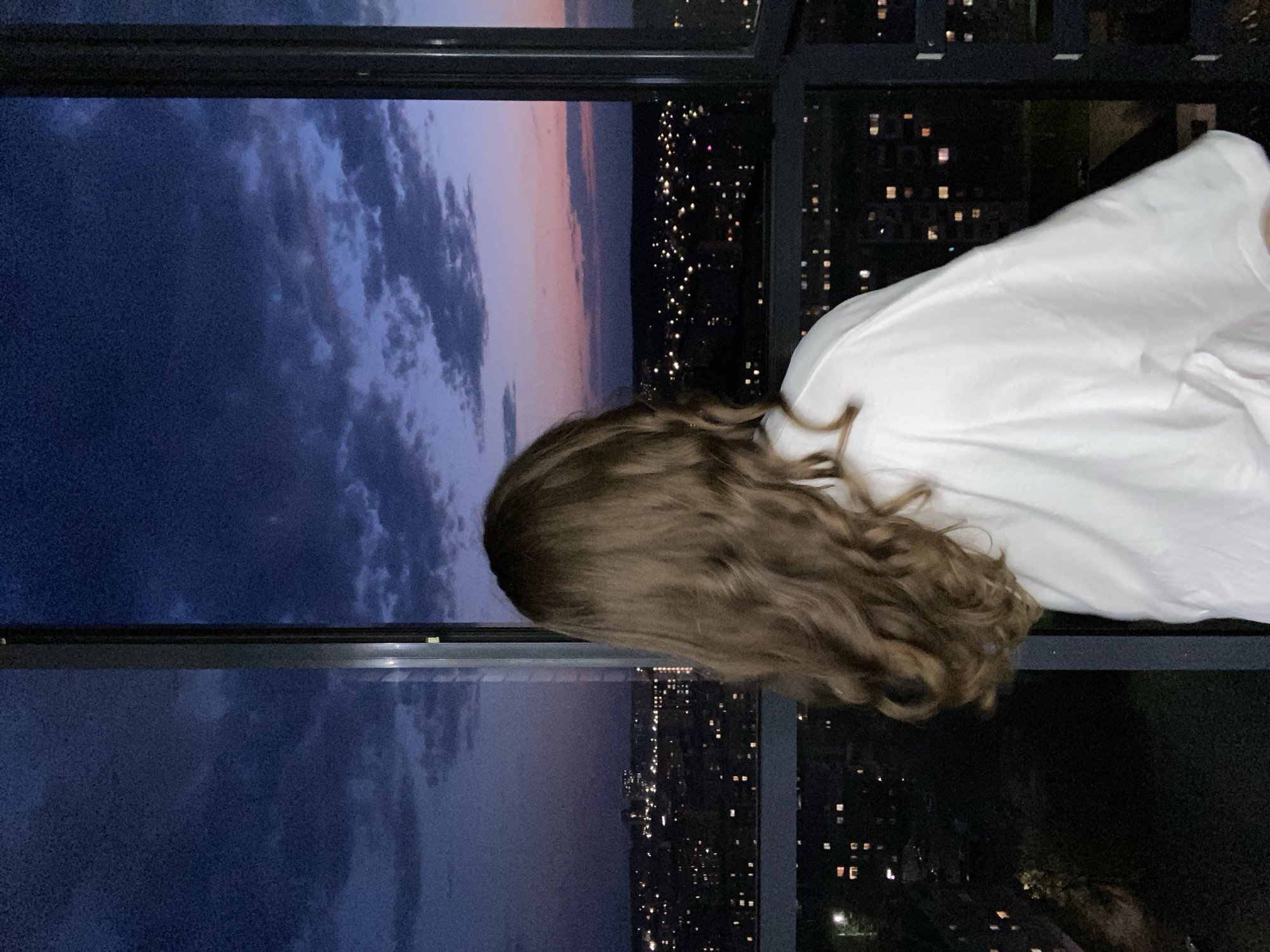 A low-angle medium shot shows a person with curly brown hair and a white shirt. The person is positioned near a multi-paned window. Outside, the night sky is transitioning from blue to pink at the horizon with scattered clouds. Below the sky, a cityscape with numerous illuminated windows stretches into the distance. The photograph was taken in Izhevsk, Russia.