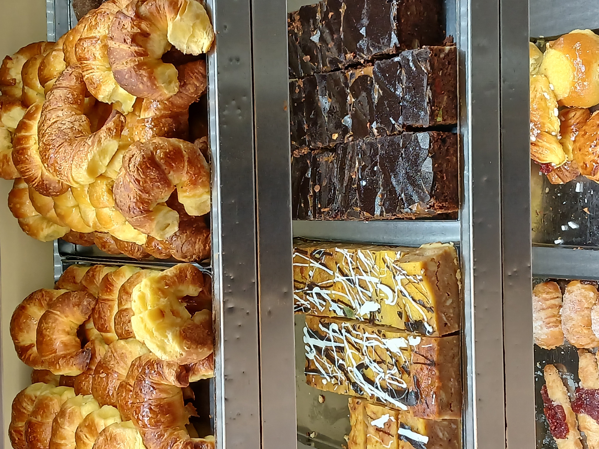 The image depicts pastries on display in a bakery, likely in Buenos Aires, Argentina, given the prevalence of medialunas (croissants) which are common there.
The display case is made of metal, with several shelves holding different types of baked goods.
On the left, there are two shelves filled with medialunas, stacked high and appearing fresh.
In the middle section, the top shelf contains rows of dark chocolate brownies. The bottom shelf has slices of a light-colored cake drizzled with white and dark chocolate.
On the right side of the image, there are buns, puff pastries with cream, and pastries with red fruit filling.