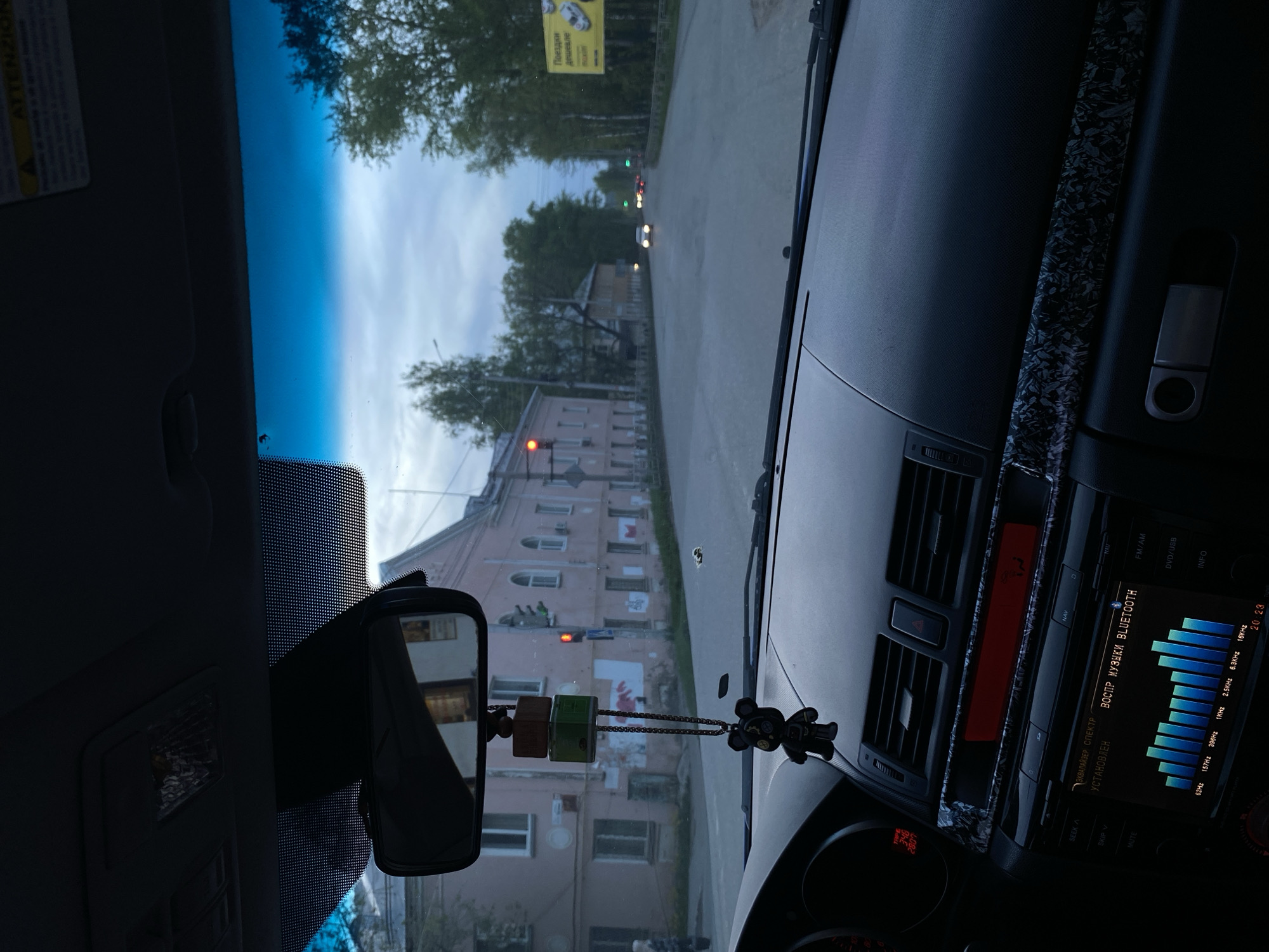 The image, taken from inside a car, shows a view through the windshield onto a street in Votkinsk, Russia. A pink brick building with multiple stories and windows is visible on the left. The car's interior is partially visible, including the rearview mirror and part of the dashboard with a display showing audio information. The display is set to Bluetooth mode, and the time is 20:23. A decorative item hangs from the rearview mirror. In the background, trees line the street, and a sign with an advertisement is visible. Traffic lights are seen in the distance.