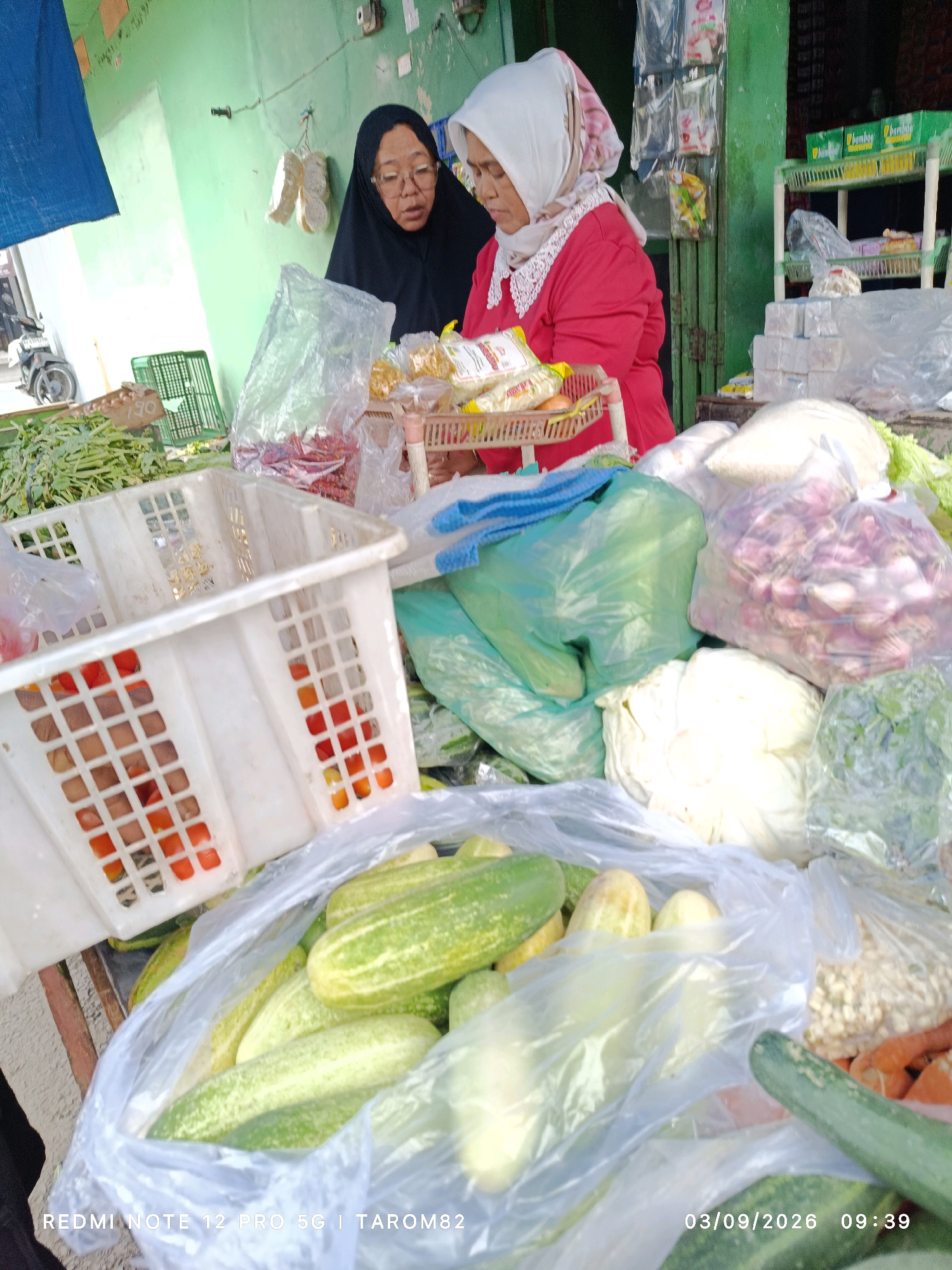 In a market stall in Kandangan, Indonesia, two women are attending to a display of fresh produce. The woman on the left wears a black hijab and glasses, while the woman on the right is dressed in a bright red top and a white headscarf. They appear to be interacting with customers or organizing the goods. The stall is laden with various vegetables, including cucumbers, cabbage, onions, and tomatoes, some of which are packaged in plastic bags and baskets. The background shows a typical market setting with shelving units and other market stalls. The lighting suggests it is daytime.