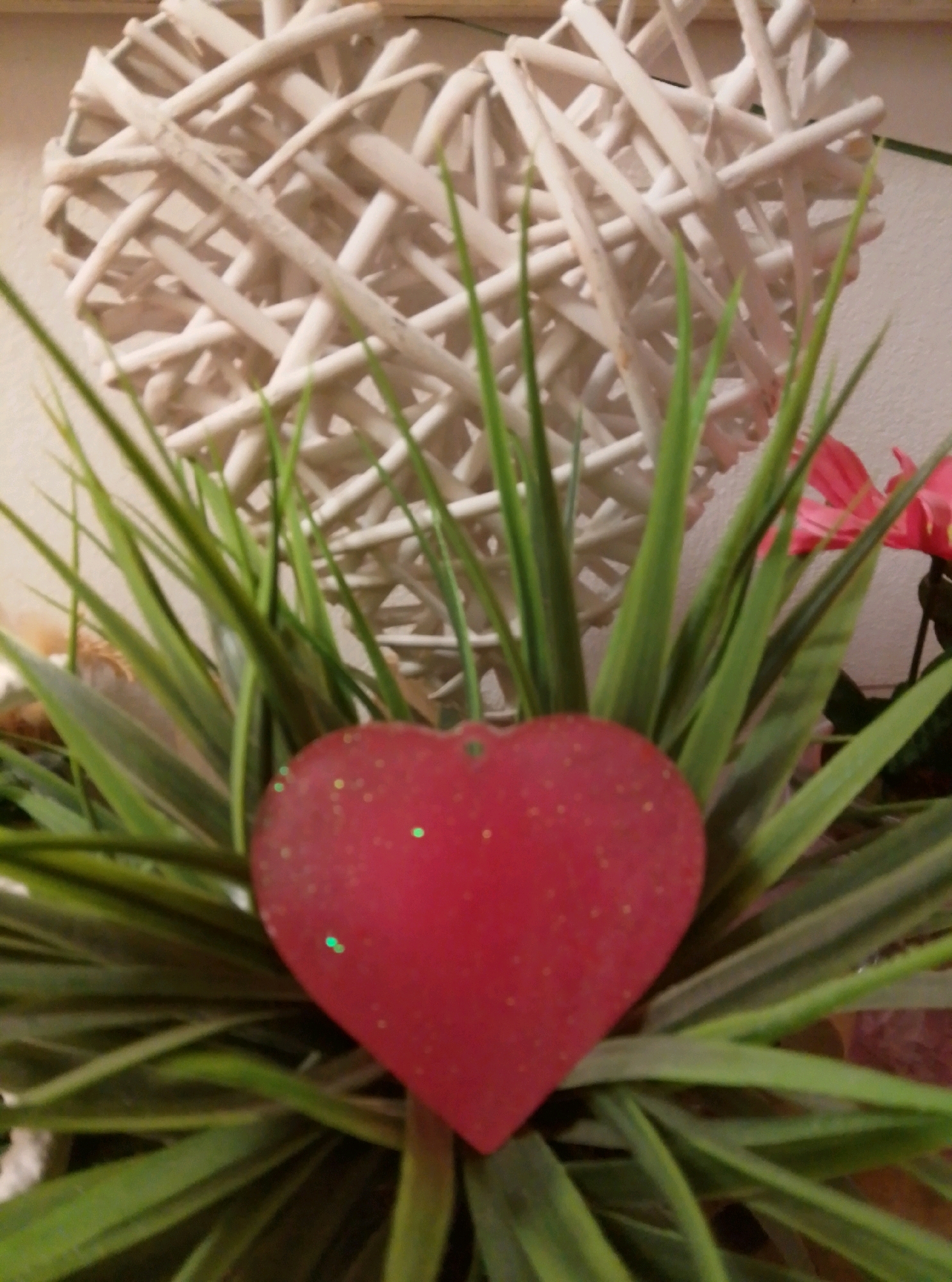 The image displays a close-up view of decorative items, likely within a residential setting. In the foreground, a red, heart-shaped ornament with glitter is positioned among vibrant green artificial grass. Behind the heart, a large, white, woven heart structure serves as a backdrop. Further back, a bright pink flower is visible. The lighting suggests an indoor environment, and the overall composition creates a festive and decorative aesthetic. There is no discernible information regarding the city of Wedel, Germany, or any individuals present in the image.