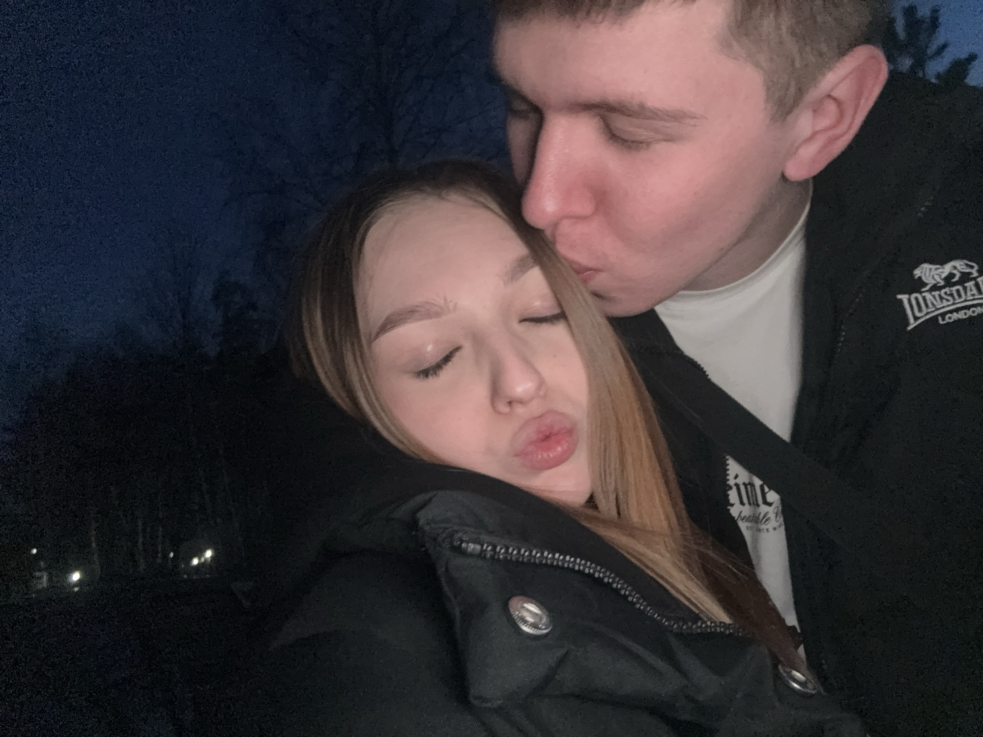A photograph shows a young man kissing a young woman on the forehead. The man wears a black jacket with the Lonsdale London logo on the right breast. Underneath the jacket, he wears a white t-shirt. The woman wears a black puffer jacket. Both have their eyes closed. The photograph appears to be taken at night, in an outdoor location with visible trees and lights in the background. The location is Chaikovsky, Russia.