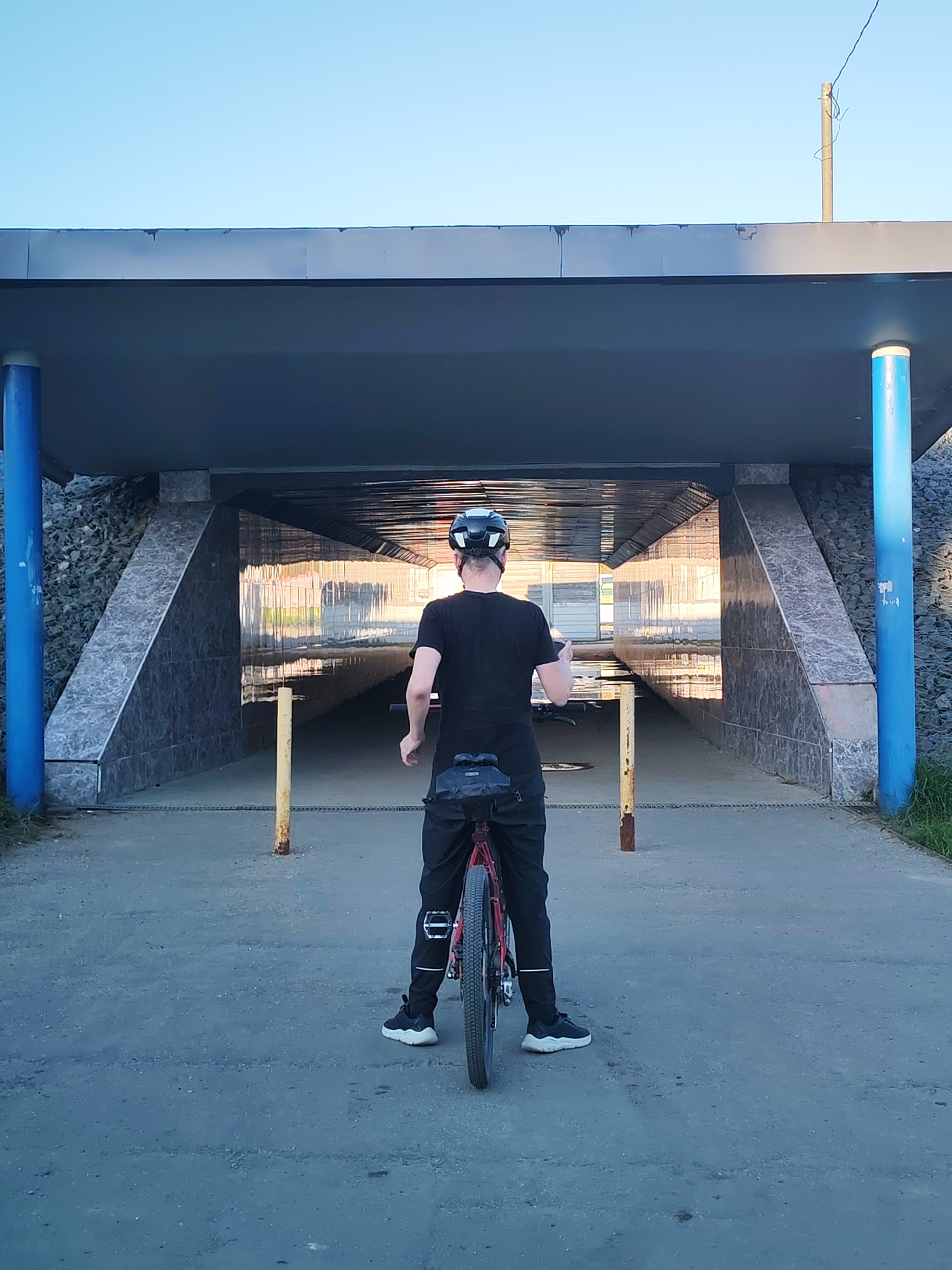 A man in a black T-shirt, dark pants, and a cycling helmet stands with his bicycle facing away from the camera, positioned in front of an underpass in Yakhroma, Russia. The underpass is gray and features blue support columns on either side of the entrance. The walls are tiled with gray marble-like stone. A rusted yellow barrier is placed on either side of the underpass entrance. The man appears to be holding a mobile phone in his hand. The sky is clear and light blue.