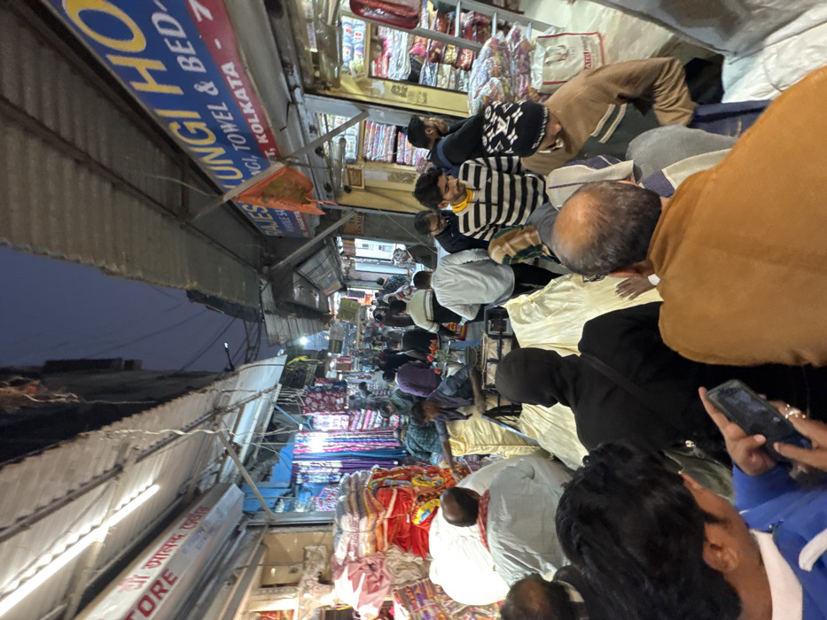 This image presents a crowded, narrow market lane in Kolkata, India, specializing in textiles. The location is identified by signage, including "RAJESH LUNGI HOUSE" which advertises "LUNGI, TOWEL & BED SHEET WHOLESALE" and explicitly mentions "KOLKATA." Another visible sign reads "श्री आलोक स्टोर."
The scene is bustling with numerous individuals, primarily adult males, navigating the congested space. Many are dressed in casual wear, including sweaters and jackets, indicative of cooler weather. In the immediate foreground, an individual holds a smartphone, seemingly capturing the scene. A person centrally positioned wears a black balaclava-like head covering.
Both sides of the lane are lined with shops, extending corrugated metal awnings that house fluorescent tube lights, providing illumination against the twilight sky. Merchandise, consisting of large, stacked bundles of folded textiles in various colors and patterns, overflows from the shops onto the lane. A handcart, heavily laden with large bundles of light-colored fabric covered in tarpaulins, is being pushed through the crowd by a man in a purple shirt, demonstrating active commercial transport within the lane. An orange flag, possibly religious, is affixed to an awning structure near "RAJESH LUNGI HOUSE." Overhead, a dense network of electrical wires is visible. The atmosphere is one of active commerce and pedestrian movement.