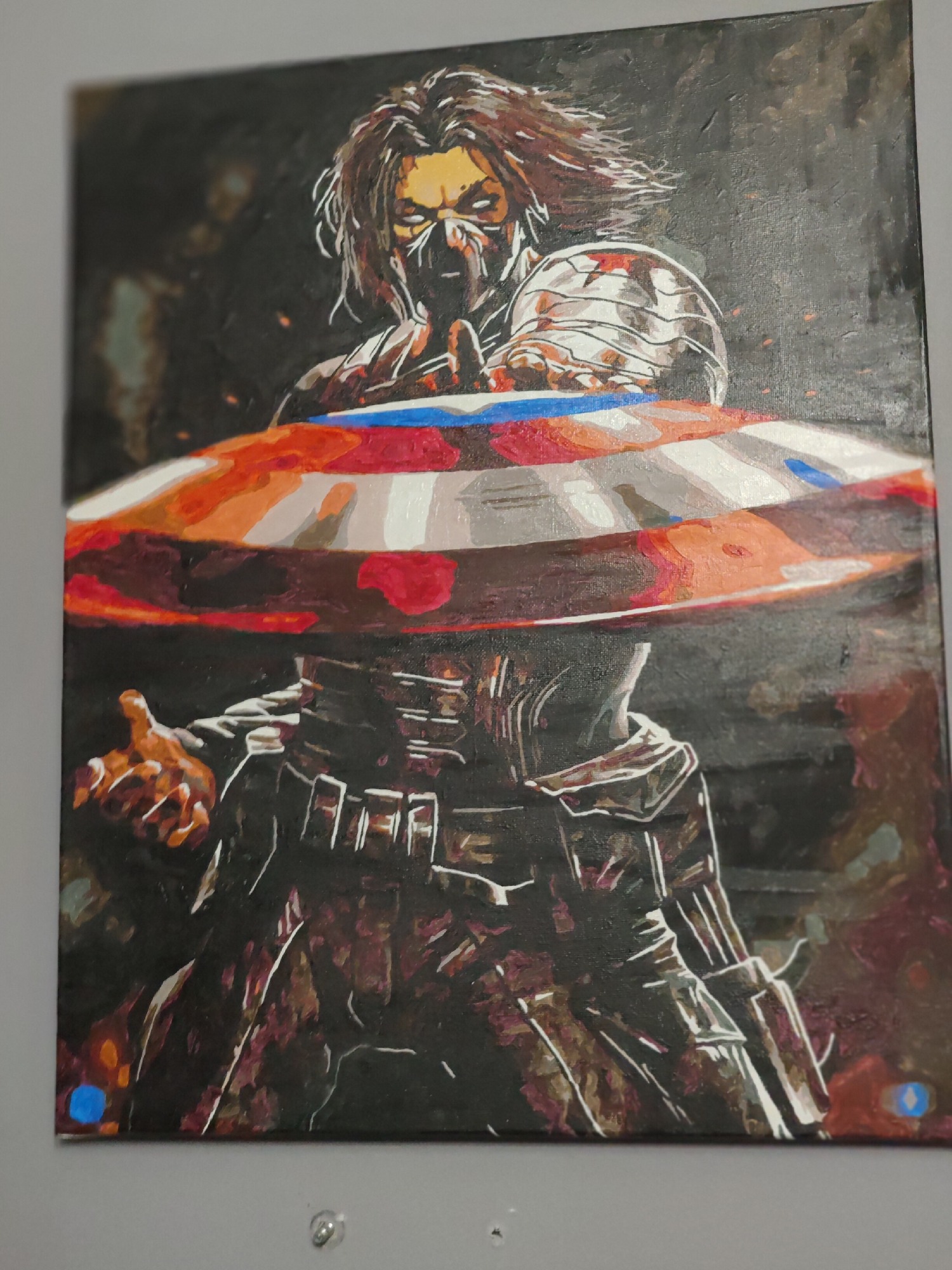An unframed, vertical, acrylic painting on canvas depicts a stylized, aggressive portrayal of the Winter Soldier from Marvel Comics. The figure is centrally positioned, with a dark, textured background that features muted orange and brown hues, possibly representing embers or distant explosions. The Winter Soldier is shown in a dynamic pose, holding Captain America's iconic shield, which dominates the foreground.
The shield is rendered with broad strokes of red, white, and blue, with segments of orange and brown suggesting wear or intense action. The Winter Soldier's upper body is clad in metallic armor, primarily depicted in silver and white, with a red star insignia visible on his shoulder. His face is partially obscured by a dark mask or balaclava, with harsh yellow and white highlights emphasizing his eyes, giving him a menacing expression. His long, dark hair appears windswept.
Below the shield, the Winter Soldier's torso is covered in a dark, armored suit, with white outlines and highlights suggesting the texture and form of the costume. One of his metal arms is bent, and his other hand is outstretched, palm facing upwards, as if to catch or deflect something. The lower part of the painting is dark, with touches of red and purple, and at the bottom left corner, two small, abstract blue shapes are visible, possibly a signature or a stylistic element. The painting is hung on a plain, light gray wall.
