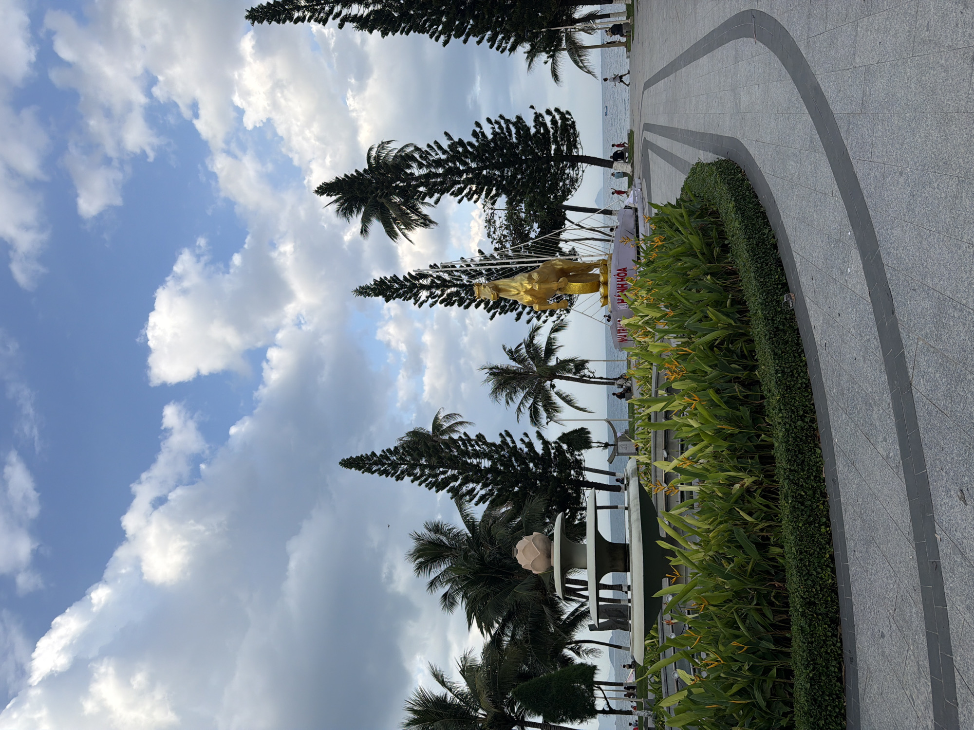 This image captures a vibrant daytime scene along a coastal promenade in Nha Trang, Vietnam. The sky is a bright blue, dotted with scattered white cumulus clouds, indicating pleasant weather.
In the foreground, a paved walkway, detailed with grey and dark grey curved patterns, borders a well-maintained strip of greenery. This verdant area features a neatly trimmed hedge and broader-leafed plants, some adorned with yellow and orange flowers, possibly bird of paradise.
Central to the composition is a striking golden statue of a horse, prominently displayed on a white, boat-like structure. An intricate white framework arches behind the statue, enhancing its presentation. Lining the promenade are various tall trees, including slender pine-like trees and several tropical palm trees.
Beyond the trees and the promenade, the calm blue expanse of the sea stretches into the distance. Several people are visible along the walkway and by the water's edge, engaging in casual leisure activities such as walking or standing, enjoying the seaside environment.
Visible text on the white base of the golden horse statue clearly reads "NHA TRANG" and "KHÁNH HÒA" in red letters, confirming the location. The scene portrays a sunny, peaceful day in a scenic coastal area, combining natural beauty with a touch of local art and public space.