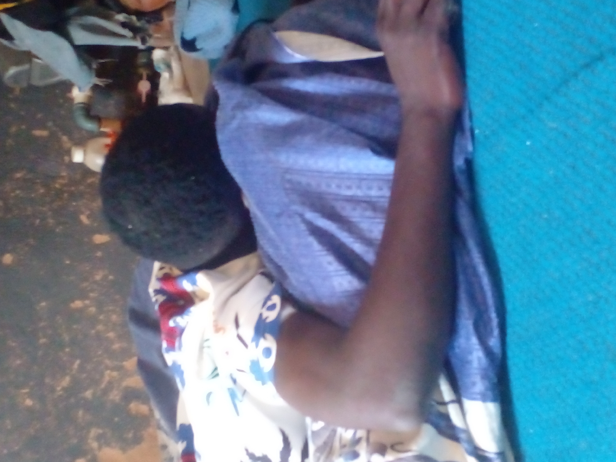 The image shows a person lying on a bed, with their head turned away from the camera. The person is wearing a blue garment. There are blankets and pillows on the bed, with one pillow having a floral pattern. The background is a textured, dirty wall. The scene appears to be indoors.