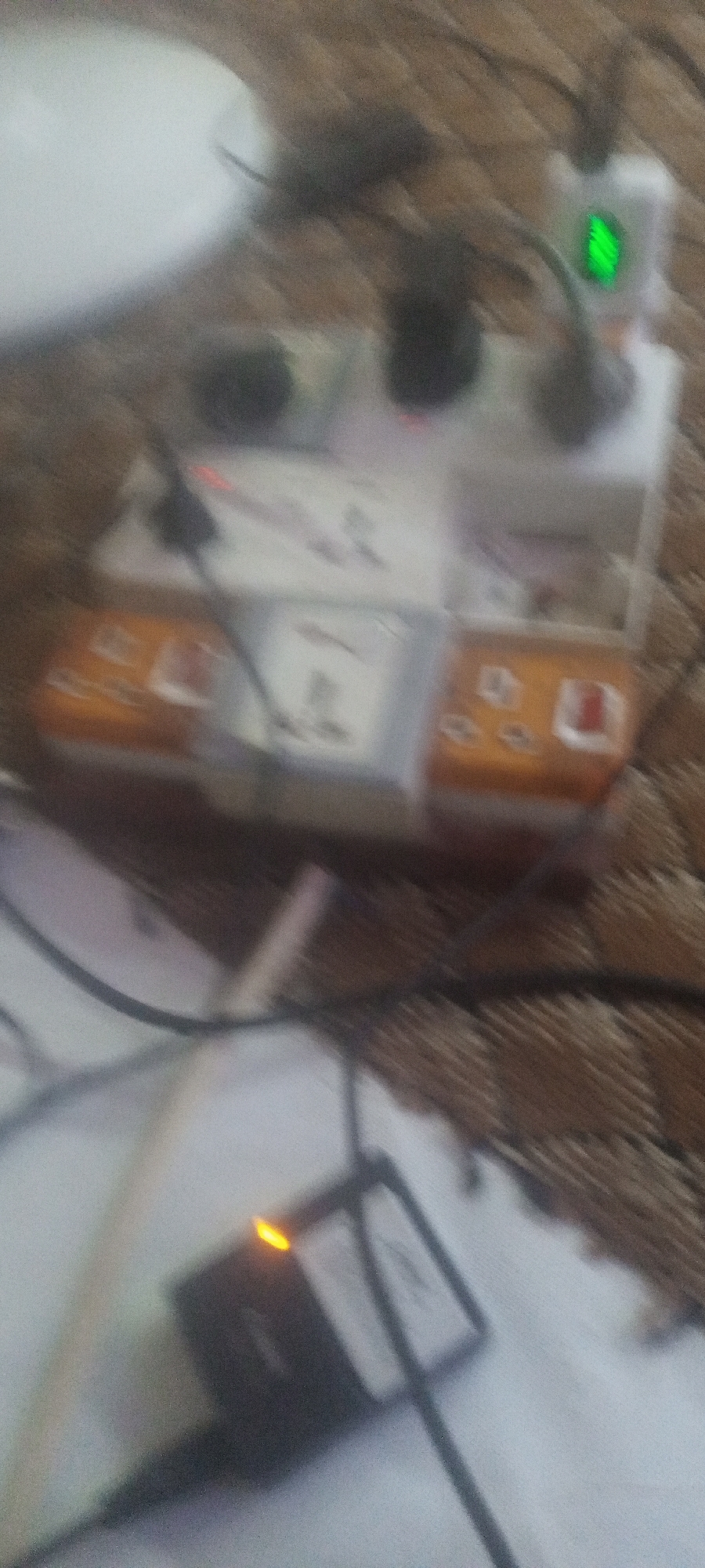 The image shows an electrical power strip with several plugs inserted. A charger with an orange light is visible in the foreground, lying on what appears to be a white surface. A white plate is visible in the background, along with a table with a patterned cloth on it. A device with a green light is plugged into the power strip. The location is unknown.