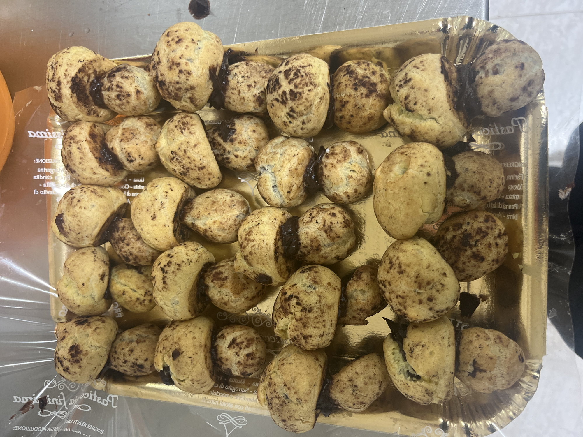 A tray of chocolate-filled cream puffs is presented on a golden tray. The cream puffs are small, round, and have a golden-brown exterior, speckled with dark brown chocolate pieces. Some of the cream puffs are split open, revealing a rich, dark chocolate filling. The tray is placed on a metallic surface, possibly a countertop. The background shows a transparent plastic wrapper and some blurred orange object. The setting appears to be a commercial kitchen or bakery. There are no people visible in the image. The text on the plastic wrapper suggests a connection to Vallo della Lucania, Italy, possibly indicating the origin or sale of these pastries.