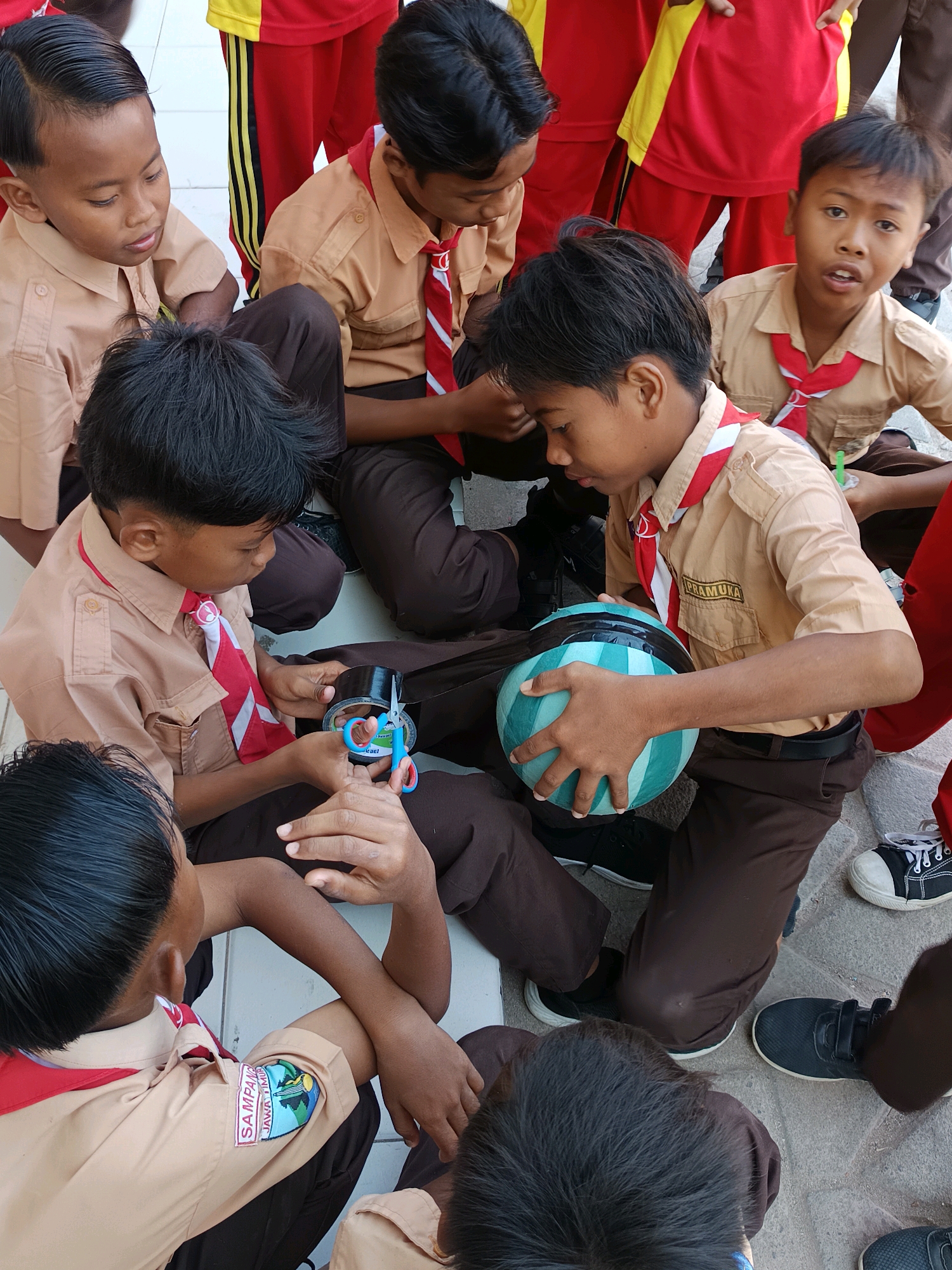 In the city of Labuhan, Indonesia, a group of young boys in scout uniforms are gathered on a paved outdoor area. They are dressed in light brown shirts with red and white neckerchiefs, and dark brown pants. Their attention is focused on a light blue and green striped ball. One boy is holding the ball, while another is using scissors and tape, suggesting they are engaged in some sort of craft or game. The background shows parts of red and yellow uniforms, possibly of other students or teachers. The overall atmosphere appears to be one of activity and collaboration.