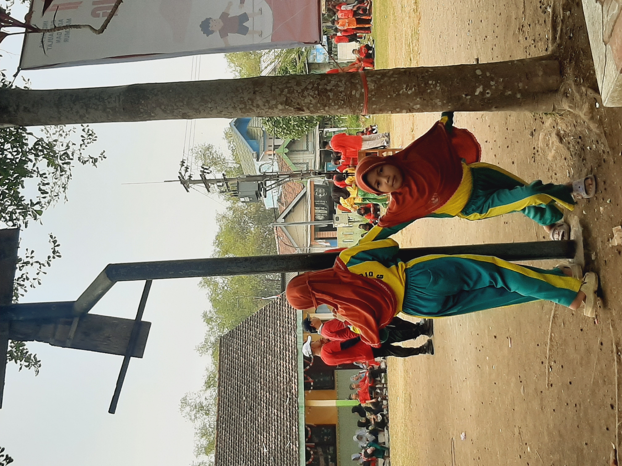The image depicts an outdoor scene in Sumberjo, Indonesia, likely a schoolyard or community space. In the foreground, two young girls are positioned near a large tree. The girl on the left, wearing a yellow and teal tracksuit with a red headscarf, is leaning against a wooden structure. To her right, another girl, also in a yellow and teal tracksuit and red headscarf, is facing the camera with her left arm extended. Behind them, a group of adults in red shirts and black pants, some wearing caps, are standing. Further in the background, there are buildings with tiled roofs and more people, some seated and others standing. A large banner with Indonesian text and illustrations is visible on the right side of the frame. The ground is covered in dirt and scattered debris. A tall pole with what appears to be electrical wires is also present. The overall atmosphere suggests an event or gathering taking place under a bright sky.