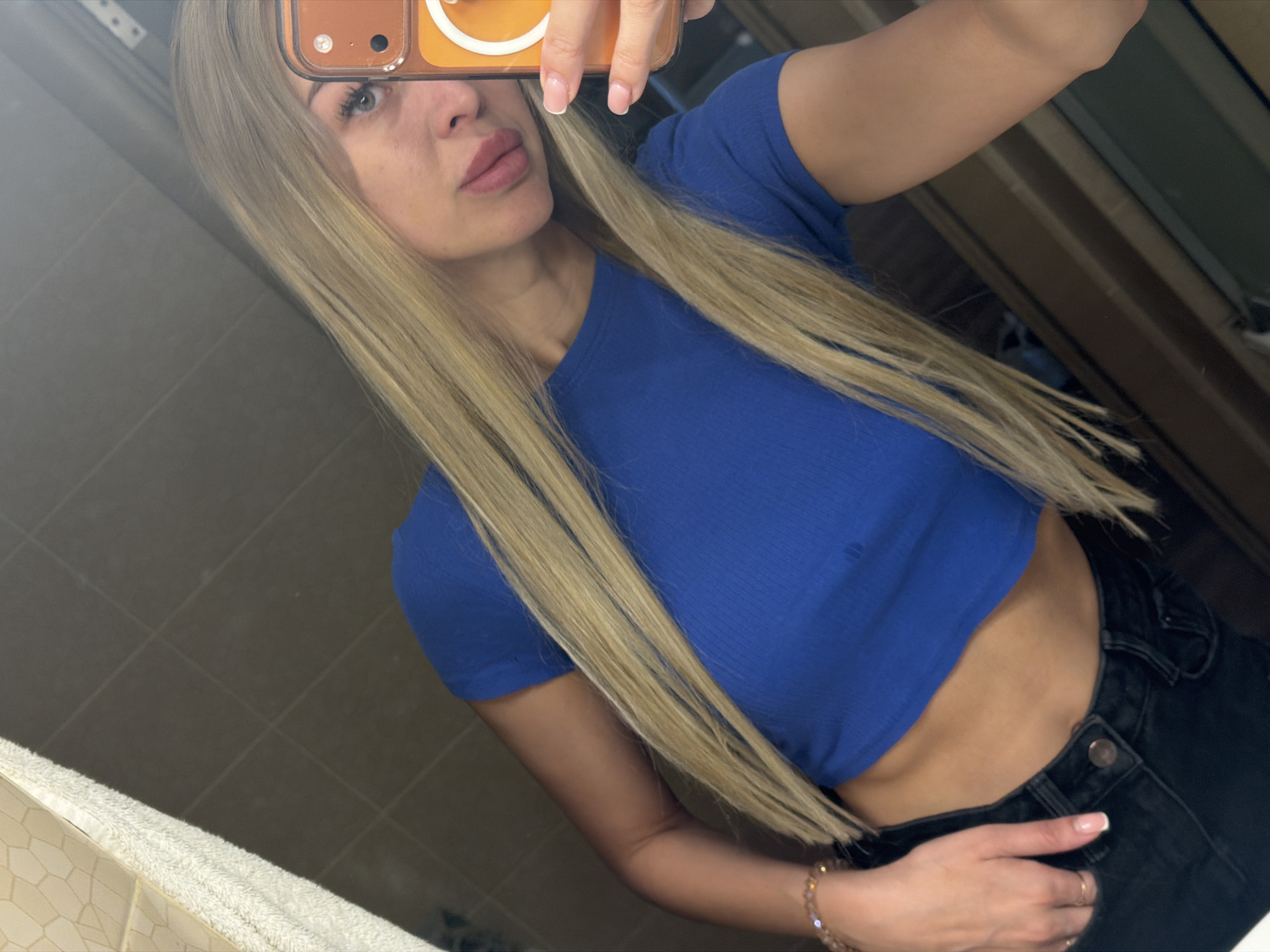 A young woman with long blonde hair takes a selfie in a dimly lit bathroom, likely in Kyiv, Ukraine. She is wearing a bright blue crop top and black jeans. Her left hand is resting on her hip, and she is holding a phone with an orange case in her right hand. The background shows tiled walls and a mirror reflecting the scene.
