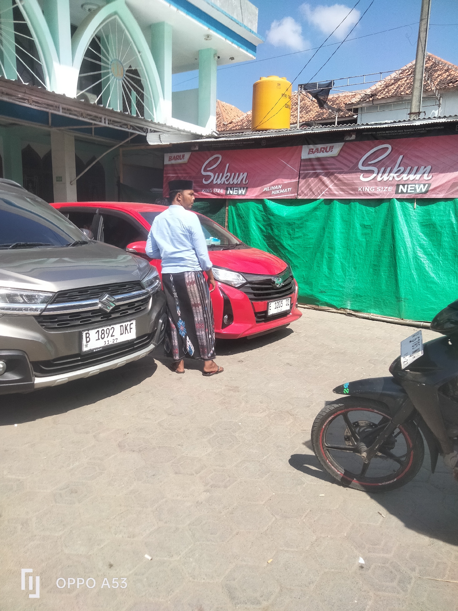 A man wearing a light blue shirt and a sarong stands next to a grey Suzuki car, facing a red car. The man is standing on a paved surface in what appears to be an outdoor area. Behind the cars, a large green tarp is visible, stretched across what might be a market stall or a temporary structure. Above the tarp, a red banner with white text reads "BARU!" and "Sukun KING SIZE NEW". Further back, the facade of a building with light blue accents and arched windows is visible, suggesting it could be a mosque or a public building. A yellow water tank is also present on the roof of a nearby structure. The sky is clear and blue, indicating a sunny day. A black motorcycle is parked in the foreground, partially obscuring the right side of the image. The license plate of the grey car reads "B 1892 DKF" and the license plate of the red car reads "E 1205 QT". The scene appears to be in a semi-urban or commercial area.