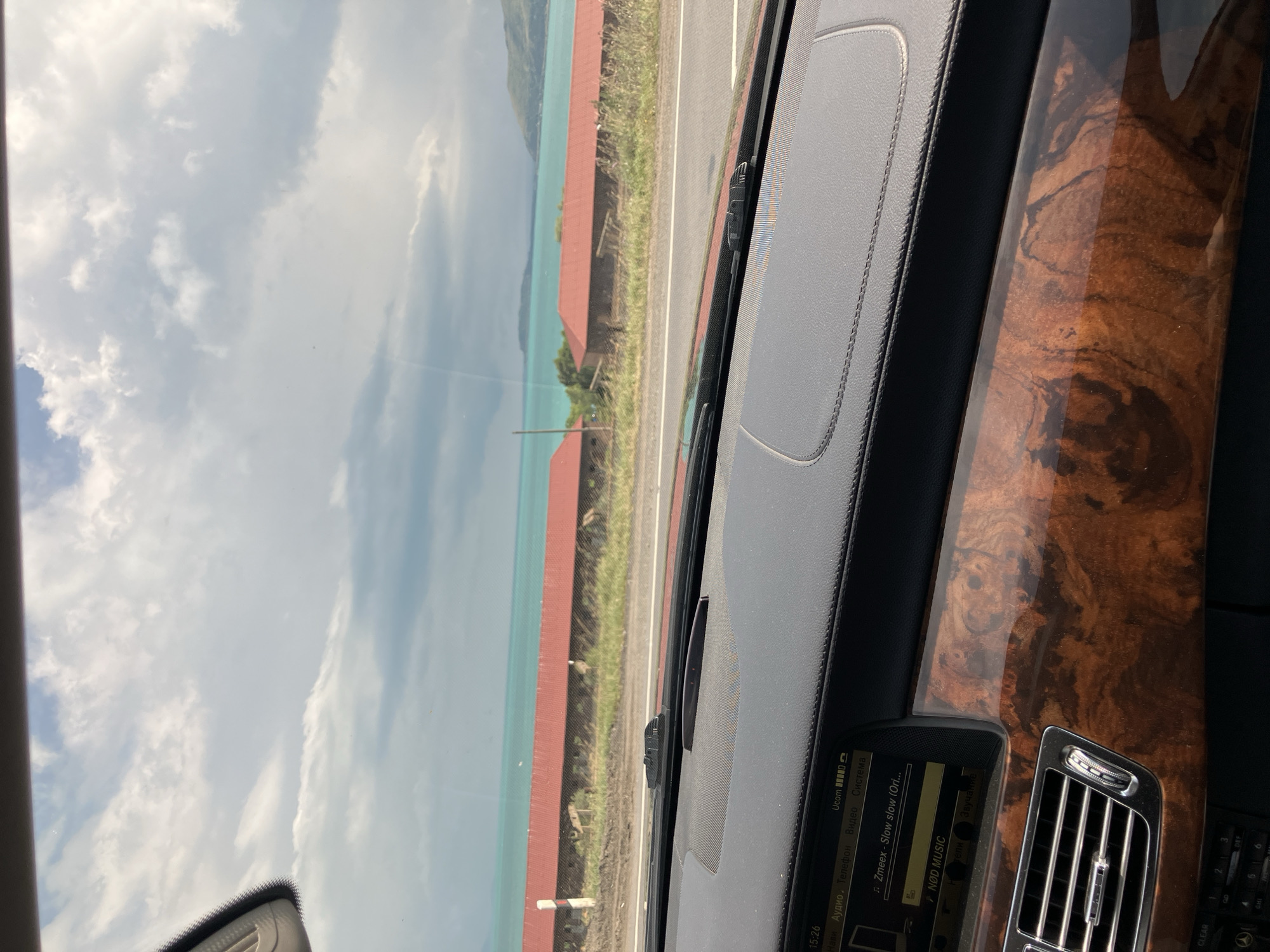 The image depicts a view from inside a car, looking out through the windshield. The dashboard is visible in the foreground, featuring a wood-grain trim and a central infotainment screen displaying music information. Outside the car, a road runs parallel to a body of water, likely Lake Sevan, given the context of Tsovagyugh, Armenia. The water is a vibrant turquoise color, and across from it, two red-roofed buildings are situated on a slight incline. The sky above is partly cloudy, with patches of blue sky interspersed with grey clouds. The overall scene suggests a scenic drive along the coast of the lake.