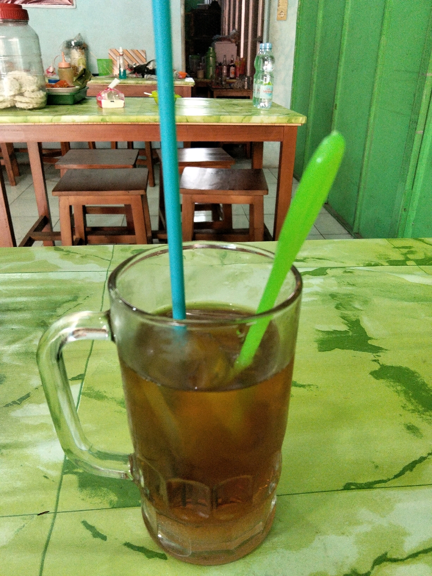 A glass of iced tea with two straws, one blue and one green, sits on a green, textured table in Panggang, Indonesia. The table is set with a wooden chair and a wooden table with a checkered tablecloth in the background. There is a large plastic bottle with crackers or biscuits inside, and some food items on the table. A green door is visible in the background. The lighting suggests it is daytime.