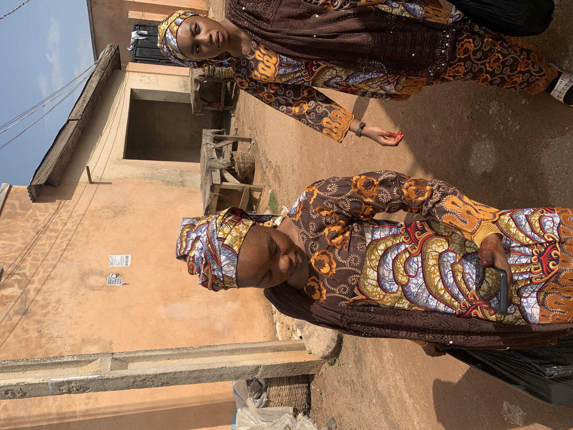 Two young women dressed in matching brown and colorful patterned traditional Nigerian attire walk on a dirt path in front of peach-colored buildings. The woman in the foreground, on the left, is looking down at a smartphone in her hand, and carrying a black plastic bag in her left hand. The woman on the right is looking to her left, with her right hand resting on her hip. In the background, there are wooden tables set up outside what appears to be a marketplace, and a clear blue sky with some power lines overhead. The scene is illuminated by bright sunlight, casting shadows on the ground. The location is identified as the city of Jos, Nigeria.