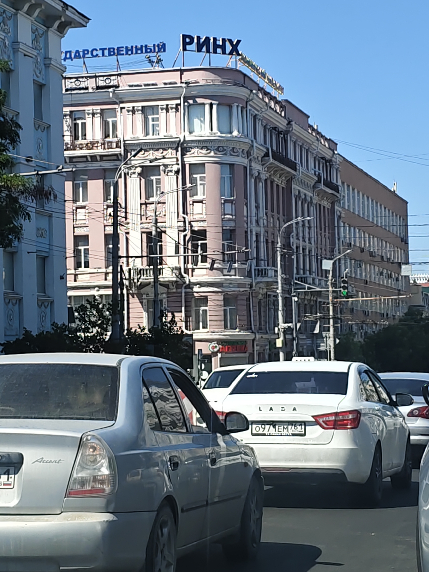 The image depicts a street scene in Rostov-on-Don, Russia, on a clear, sunny day. In the foreground, a silver Hyundai Accent is stopped in traffic, facing away from the camera. Its license plate is partially visible, showing "934 RS". To the right of the Accent, a white Lada sedan is also stopped in traffic, with its license plate reading "0971EM 761". Behind the Lada, another white vehicle is partially visible.
The middle ground features a multi-story building with a distinctive pink facade and ornate architectural details, including balconies and decorative moldings. A blue sign on the building reads "ДАРСТВЕННЫЙ РИНХ". Traffic lights are visible, with one showing a green light. Several utility poles with numerous wires are present.
In the background, more buildings are visible, including a longer, darker building to the right. Trees and foliage are present on the left side of the image, partially obscuring a lighter-colored building. The sky is a clear blue. The scene suggests a typical urban environment with moderate traffic congestion.