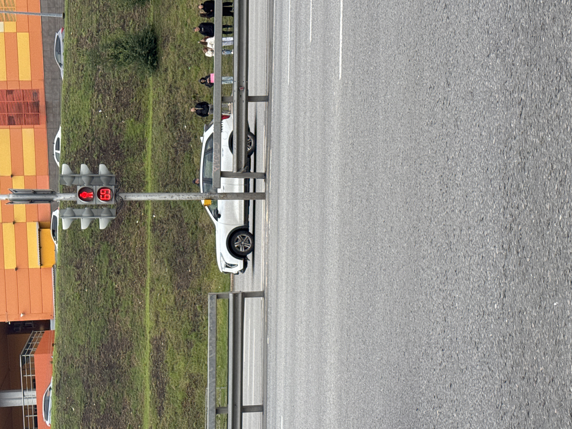 The image shows a traffic light displaying a red pedestrian signal and a red "69" in digital format, likely indicating a time remaining. In the background, a white SUV is parked on the side of the road, and several people are standing on the grassy embankment behind a metal barrier. The environment appears to be an urban setting with buildings in the far background, suggesting it could be a city like Ufa, Russia. The road is paved, and the traffic light is on a pole.