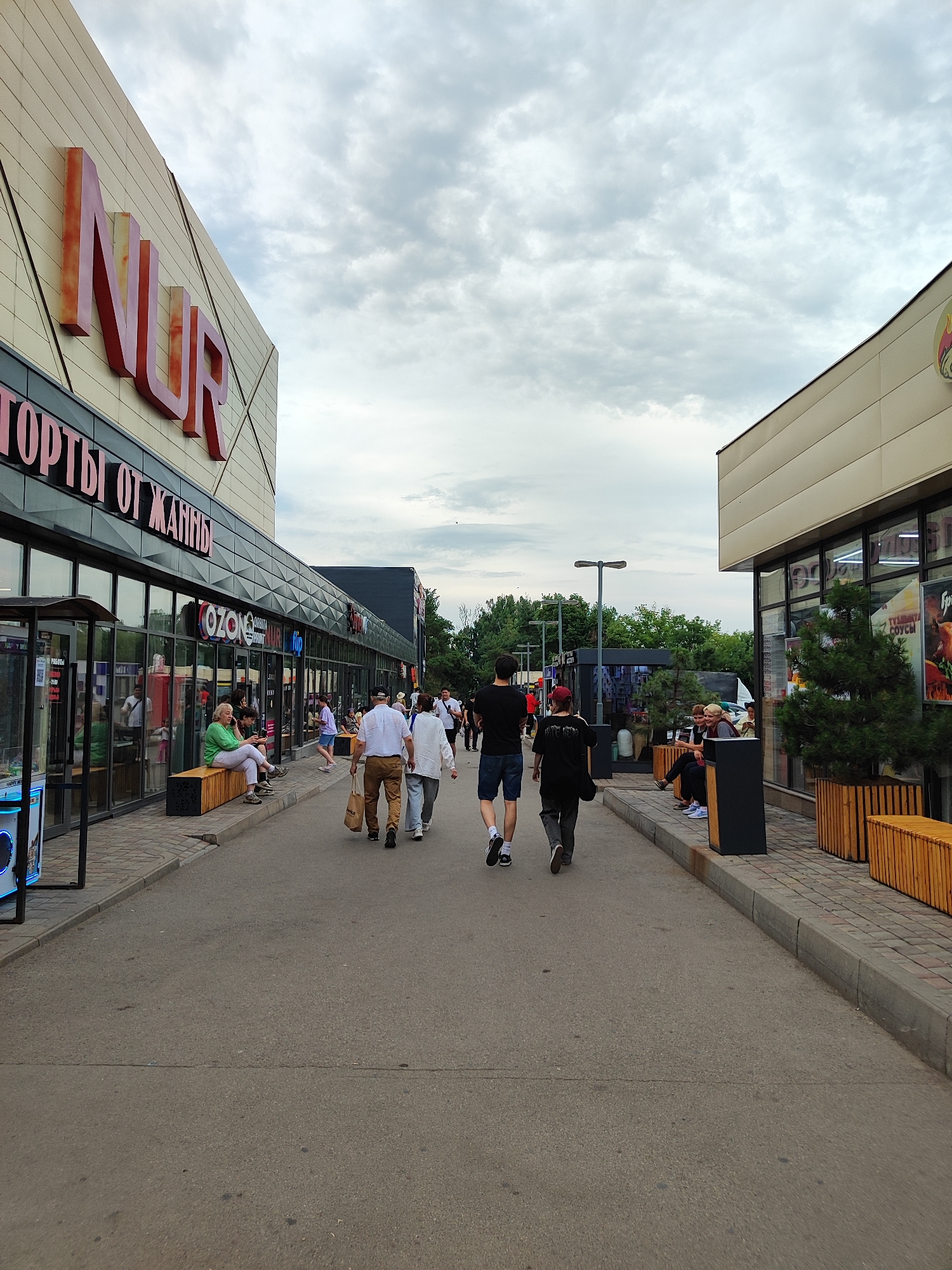 The image shows an outdoor pedestrian walkway in front of a shopping center, with several people walking and sitting. The shopping center features large windows displaying storefronts and signage. The building on the left has prominent red lettering that appears to spell "NUR". Below "NUR," there is text in Cyrillic, possibly indicating shop names or categories, such as "ТОРТЫ ОТ ЖАННЫ" (Cakes from Zhanna). Another storefront visible is "OZON."
Several individuals are captured walking away from the camera, including a man in a black t-shirt and shorts, another in a dark t-shirt and grey pants, and a couple in white shirts and light-colored pants. One woman in a green top is seated on a wooden bench, looking at her phone. Other people are also seated or standing near the storefronts.
The scene is set during the daytime, under a cloudy sky, suggesting overcast weather. The walkway is paved, with a curb and decorative wooden planters. Trees and greenery are visible in the background, indicating an urban or suburban outdoor environment. The overall atmosphere appears relaxed, with people engaged in everyday activities such as shopping or spending leisure time. The presence of multiple shops and pedestrian traffic suggests a commercial area.