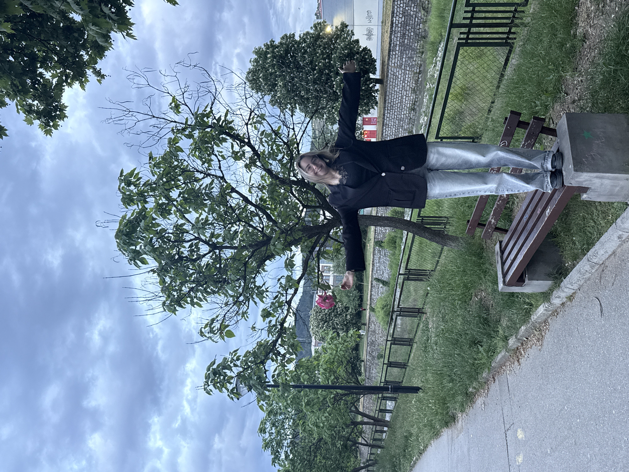 A person stands on a concrete structure beside a park bench in an outdoor area with a mix of grassy ground, trees, and a paved path. The person is wearing a dark jacket and light-colored jeans and has their arms outstretched. A tree appears behind them. The location is likely Blagoevgrad, Bulgaria.