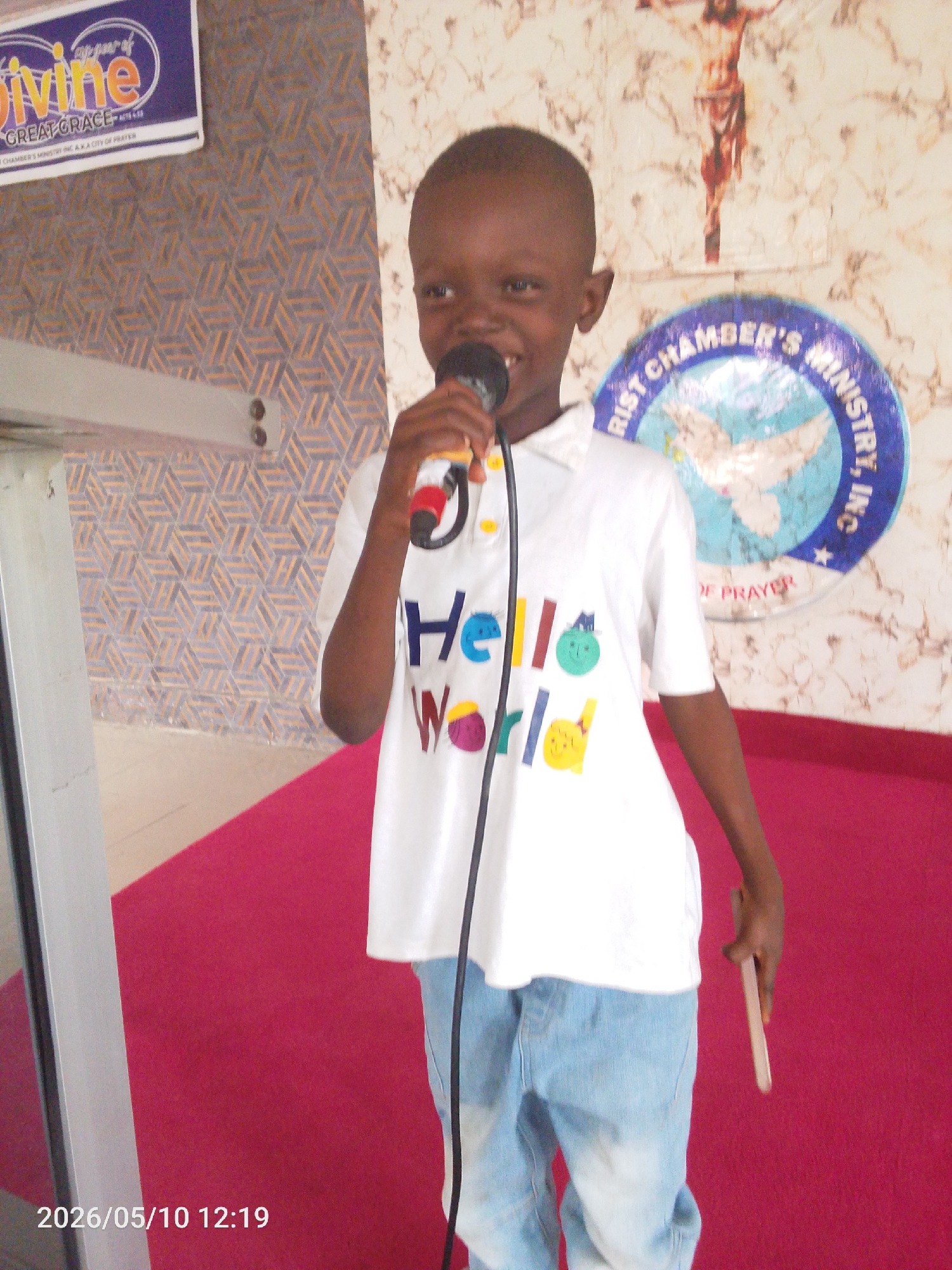 A young boy stands holding a microphone, possibly speaking or singing. He is wearing a white t-shirt with the words "Hello World" written on it and light blue jeans. In his left hand, he holds a thin, rectangular object.
The background shows a patterned wall with a crucifix and a circular emblem with a dove inside. Above the boy's head is a sign with the word "divine" visible. The location is likely a church or religious setting.
The OCR text "RIST CHAMBER'S MINISTRY, IN. DE PRAYER" suggests the setting is associated with a ministry. The OCR text "CHAMBER'S MINISTRY IN. A.K.A CITY OF PRATER" implies the location is known as the City of Prayer.
While the specific details to pinpoint the exact location to Lokoja, Nigeria, from the image is unknown, the image points toward a religious setting somewhere in the city.