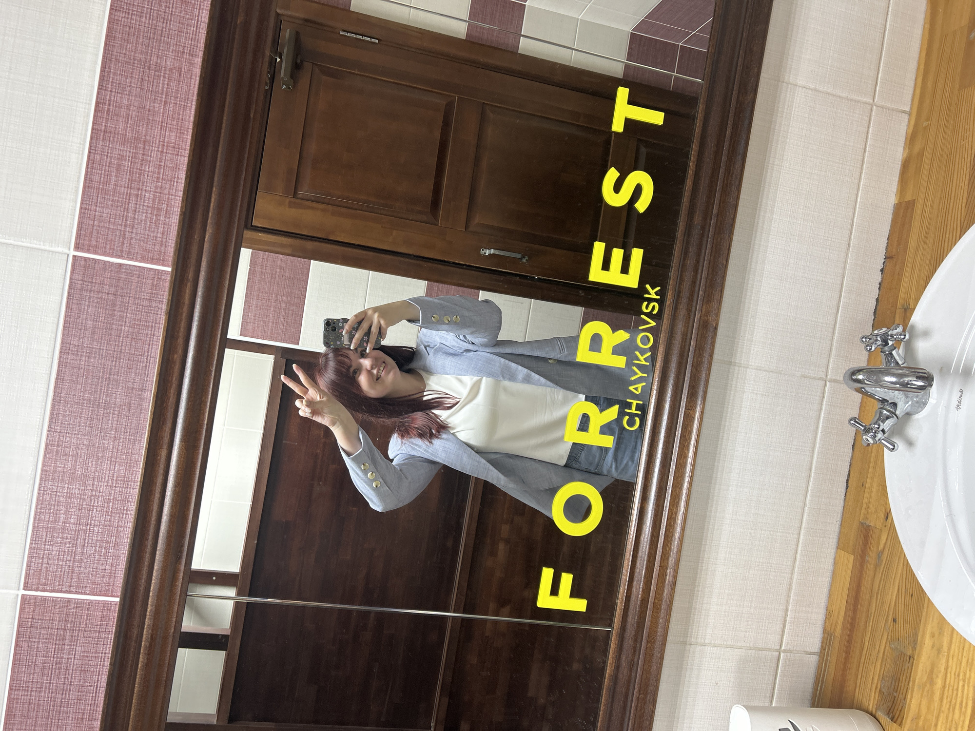 A woman is reflected in a mirror, taking a selfie in what appears to be a bathroom. The mirror has a dark wooden frame and a cabinet behind the reflection. The woman is wearing a light blue blazer over a white top. She is holding a smartphone and making a peace sign with her left hand. The text "FORREST CHAYKOVSK" is overlaid on the mirror, with "CHAYKOVSK" oriented vertically. A white sink is visible in the lower right corner, with a silver faucet. The bathroom walls have alternating rows of white and pinkish-purple tiles. The countertop to the right of the sink is made of wood.