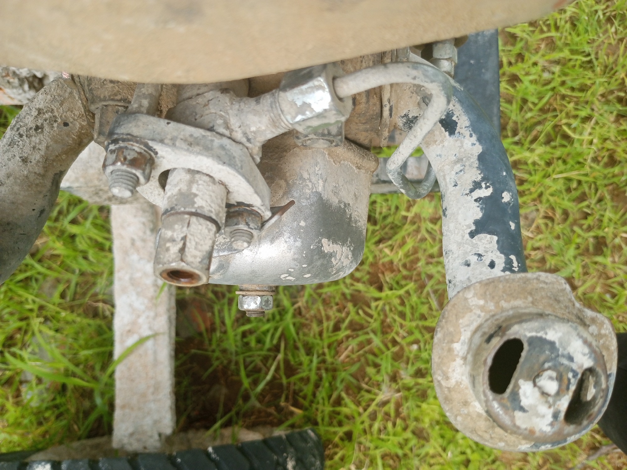 The image shows a close-up, overhead view of a dirty, metallic mechanical component, likely part of a motorcycle or similar vehicle, resting on a patch of green grass. The object appears to be a junction or mounting point, with several bolts, nuts, and pipes visible. One prominent part is a silver-colored, somewhat rounded casing with a hole in it, possibly a housing for some kind of mechanism. Another section is a black, curved pipe, also dirty and showing signs of wear. A silver bracket with two bolts is attached to a pipe on the left. The overall impression is one of outdoor exposure, given the grass and dirt. The lighting suggests it's daytime, with no clear indication of time of day or weather conditions beyond what is implied by the natural setting. There is no visible text in the image. The location context is Jalingo, Nigeria.
