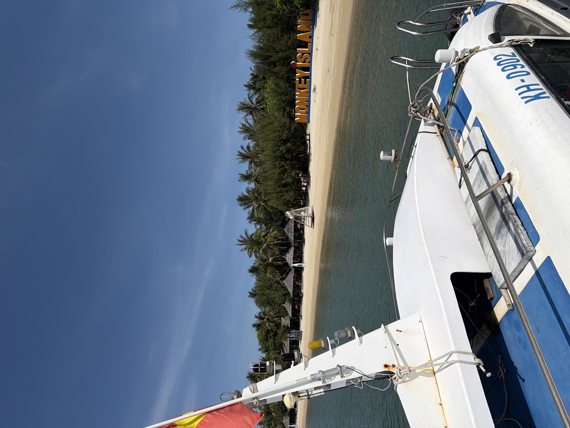 The image captures a bright, sunny day at Monkey Island, Ninh Ích, Vietnam. In the foreground, the white hull of a boat is prominently featured, with blue accents and the registration number "KH-D902" visible. Various ropes and metallic railings are attached to the boat's structure. Behind the boat, a sandy beach stretches along the shoreline, lined with palm trees and dense green foliage. Several thatched-roof huts are visible on the beach, suggesting resort accommodations. A large, orange sign with blue trim spelling out "MONKEY ISLAND" stands on the sand. The water is a calm, turquoise color, reflecting the clear blue sky. A red and yellow flag flutters from a flagpole on the boat in the upper left portion of the frame. The scene conveys a tropical island atmosphere.