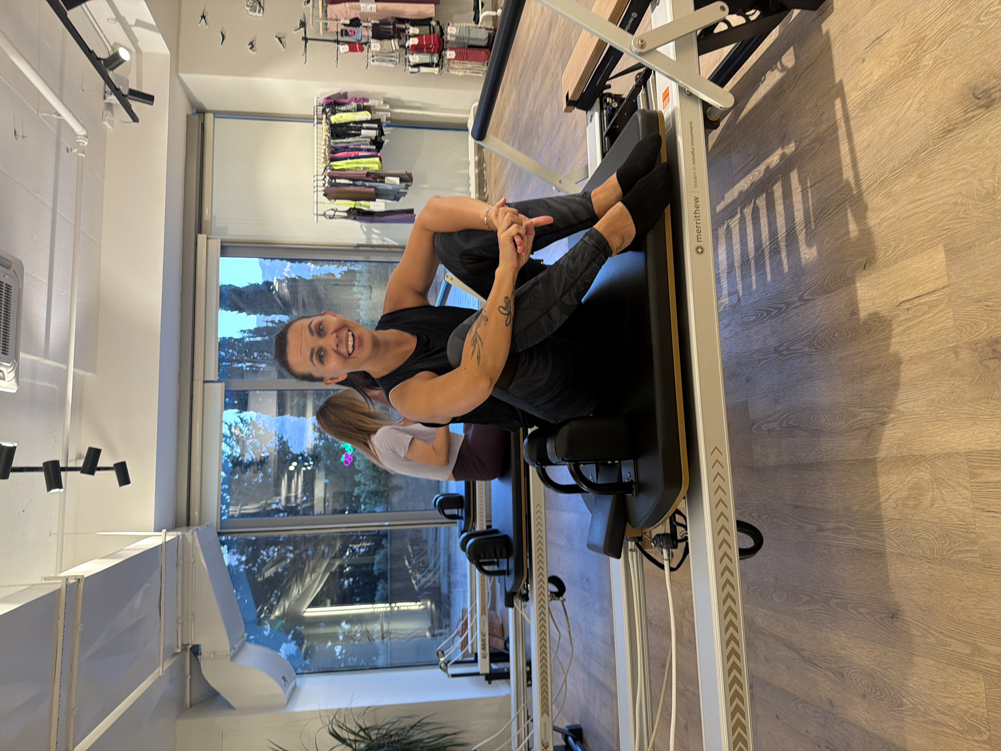 The image shows a woman sitting on a Pilates reformer in a studio. The studio appears to be located in Boreti, Montenegro, as indicated by a sign on the reformer which reads "merrithew. Leaders in mindful movement." The woman is smiling and looking at the camera. She is wearing black workout clothes and black socks. There are other Pilates reformers visible in the background, as well as a rack of workout clothes. The room has large windows that look out onto trees and a body of water. The floor is made of wood. The lighting in the room is bright and airy.