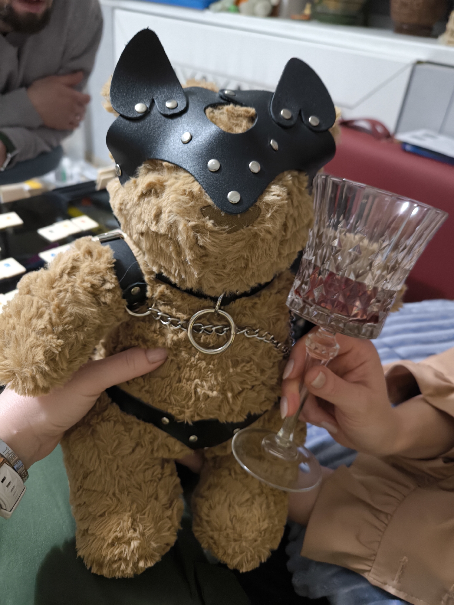 A brown teddy bear is dressed in a black leather mask with silver studs and a black leather harness with a silver metal chain, held by two people, one wearing a green shirt and a white wristwatch on their left wrist, and the other wearing a light brown shirt and holding a faceted crystal wine glass with red wine inside, indoors. The scene includes a table with black and yellow objects, a red couch, and some decorative objects on a shelf in the background. The city and specific location are not discernible.