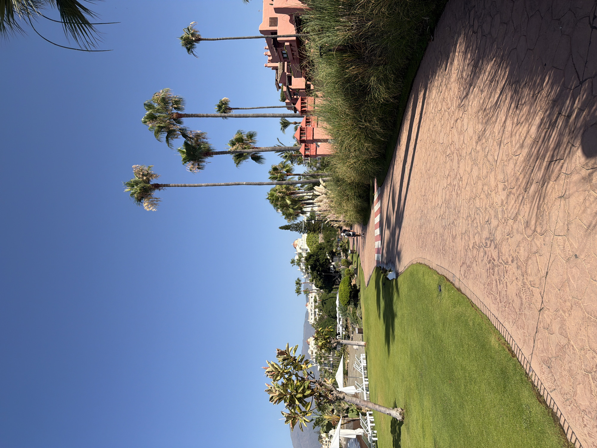This is a ground-level, full-frame, outdoor photograph taken on a sunny day in Estepona, Spain, likely along the beachfront promenade. The scene is dominated by a curved, textured, reddish-brown paved walkway that winds through the frame. To the left of the walkway is a neatly manicured green lawn with a single, slender tree casting a shadow. Further left, near the edge of the image, is a section of beach with white lounge chairs and umbrellas visible.
In the background, a line of buildings with white facades and red-tiled roofs, characteristic of Mediterranean architecture, is nestled behind lush vegetation and tall palm trees. The sky is a clear, bright blue.
On the walkway, a person wearing a backpack is walking away from the camera, towards the buildings. Another person appears further ahead on the walkway. The overall impression is one of a well-maintained resort or public area by the sea.