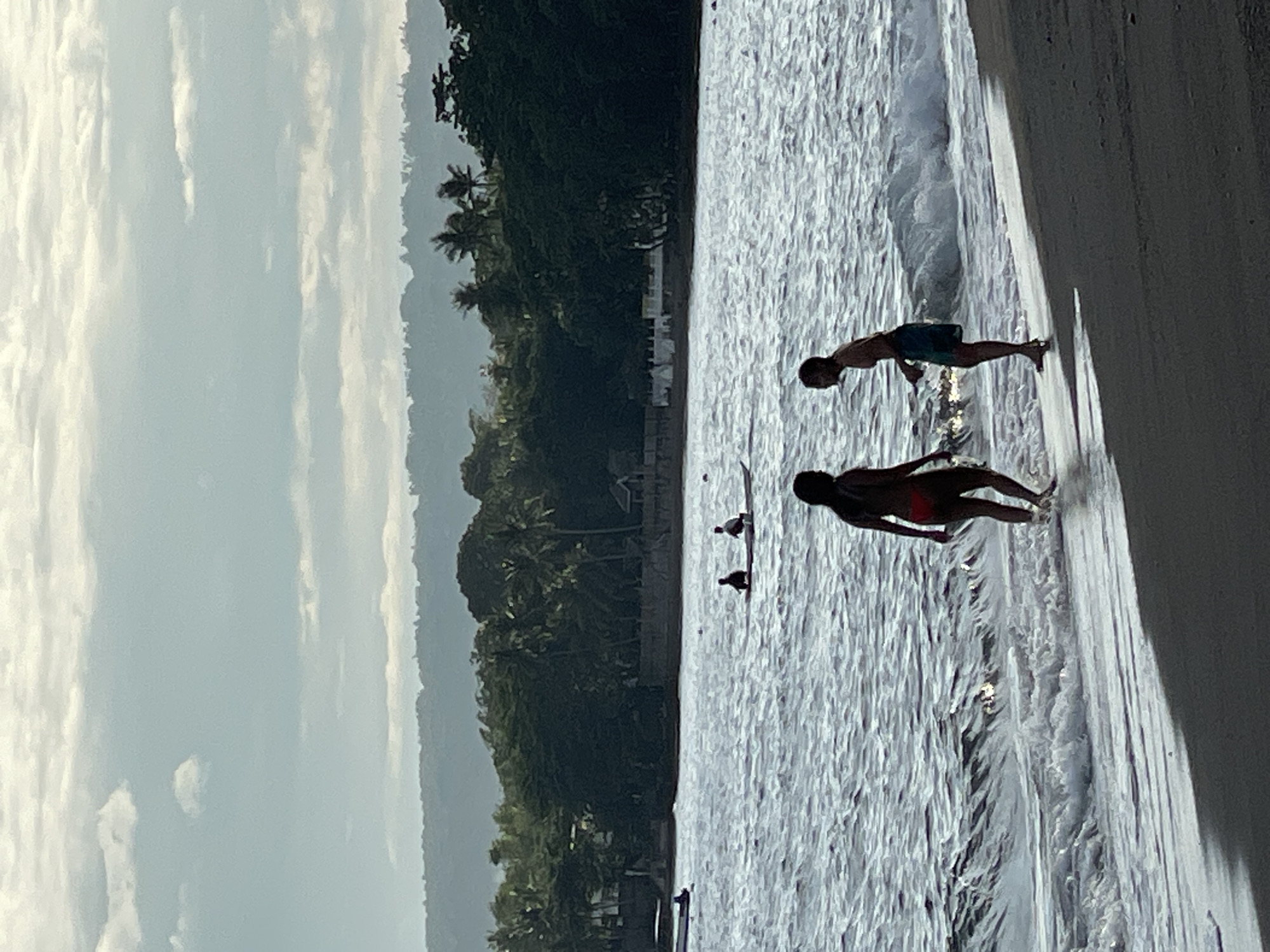 The image captures a serene beach scene in Dauin, Philippines, at dusk. In the foreground, two figures, a man and a woman, stand near the water's edge. The woman, silhouetted against the light, wears a red bikini and faces away from the camera, her body angled towards the man. The man, also silhouetted, wears blue shorts and faces the woman, appearing to be engaged in conversation or interaction with her. The ocean is calm with gentle waves lapping at the shore, reflecting the soft light of the setting sun.
Further out in the water, a long, narrow boat carries two figures, possibly fishermen, paddling through the calm sea. In the background, a dense line of lush green trees and palm trees forms a natural barrier, with a few buildings and structures visible amongst them, suggesting a coastal village or resort. The sky above is a mix of soft blues and whites, with scattered clouds indicating the transition from day to night. The overall atmosphere is peaceful and tranquil, characteristic of a tropical evening.