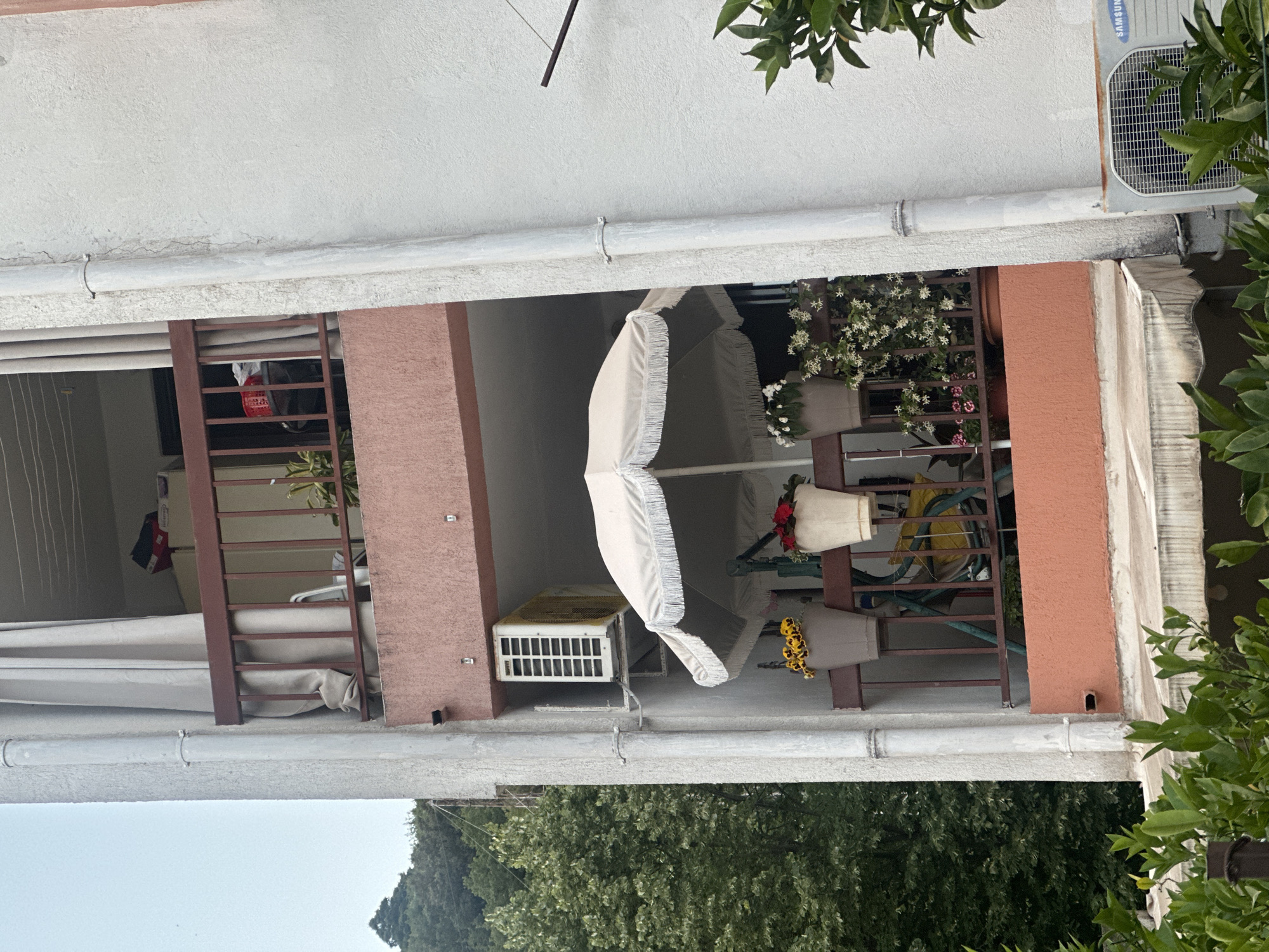 The image shows a balcony in Budva, Montenegro. The balcony has two sections, one covered by curtains and the other open. A white umbrella is positioned in the open section. There are several potted plants with flowers on the balcony. The building has a light-colored facade with some reddish-brown accents. An air conditioning unit is mounted on the building wall. Green foliage is visible in the foreground and background, suggesting the presence of trees.