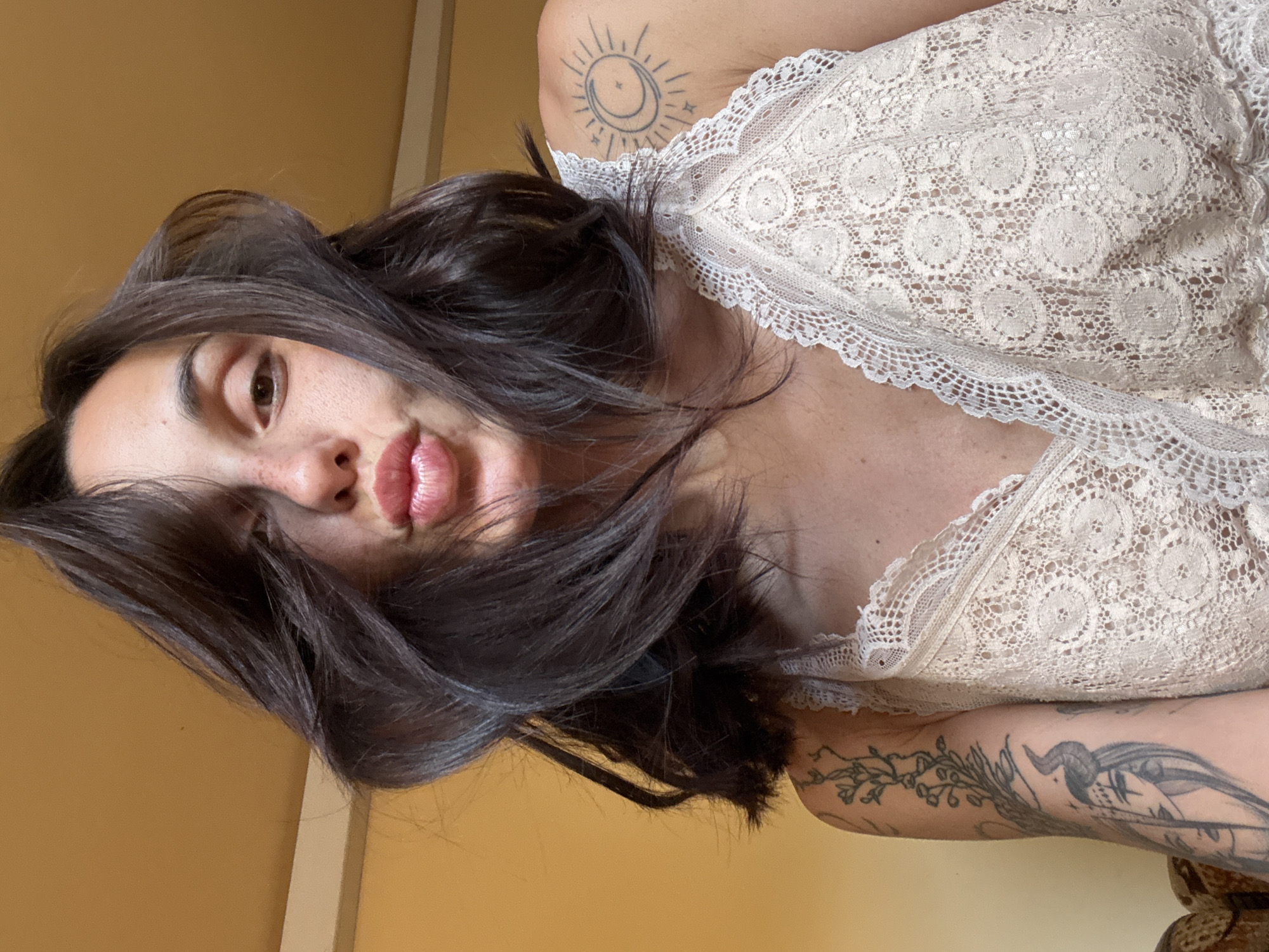 The image shows a close-up, low-angle selfie of a woman with dark hair and freckles, puckering her lips. She is wearing a cream-colored lace bralette. A tattoo of a crescent moon and sun is visible on her left shoulder, and a tattoo of a figure with horns and a crown is on her left arm. The background is a plain yellow wall. There is no indication of the city of Pleven, Bulgaria, in the image.