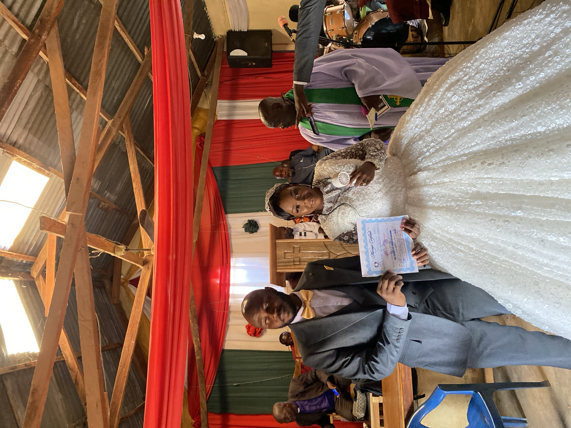 The image depicts a wedding ceremony in Vom, Nigeria. A bride in a white, beaded gown and a groom in a grey suit and gold bowtie are holding a marriage certificate. They are standing in front of red, white, and green decorations. A drummer and a man in a purple robe with a green sash stand behind them, as well as a drum set and speaker on a makeshift stage. The venue appears to be a simple structure with exposed wooden beams and a corrugated metal roof. The scene shows the couple smiling and holding the marriage certificate.