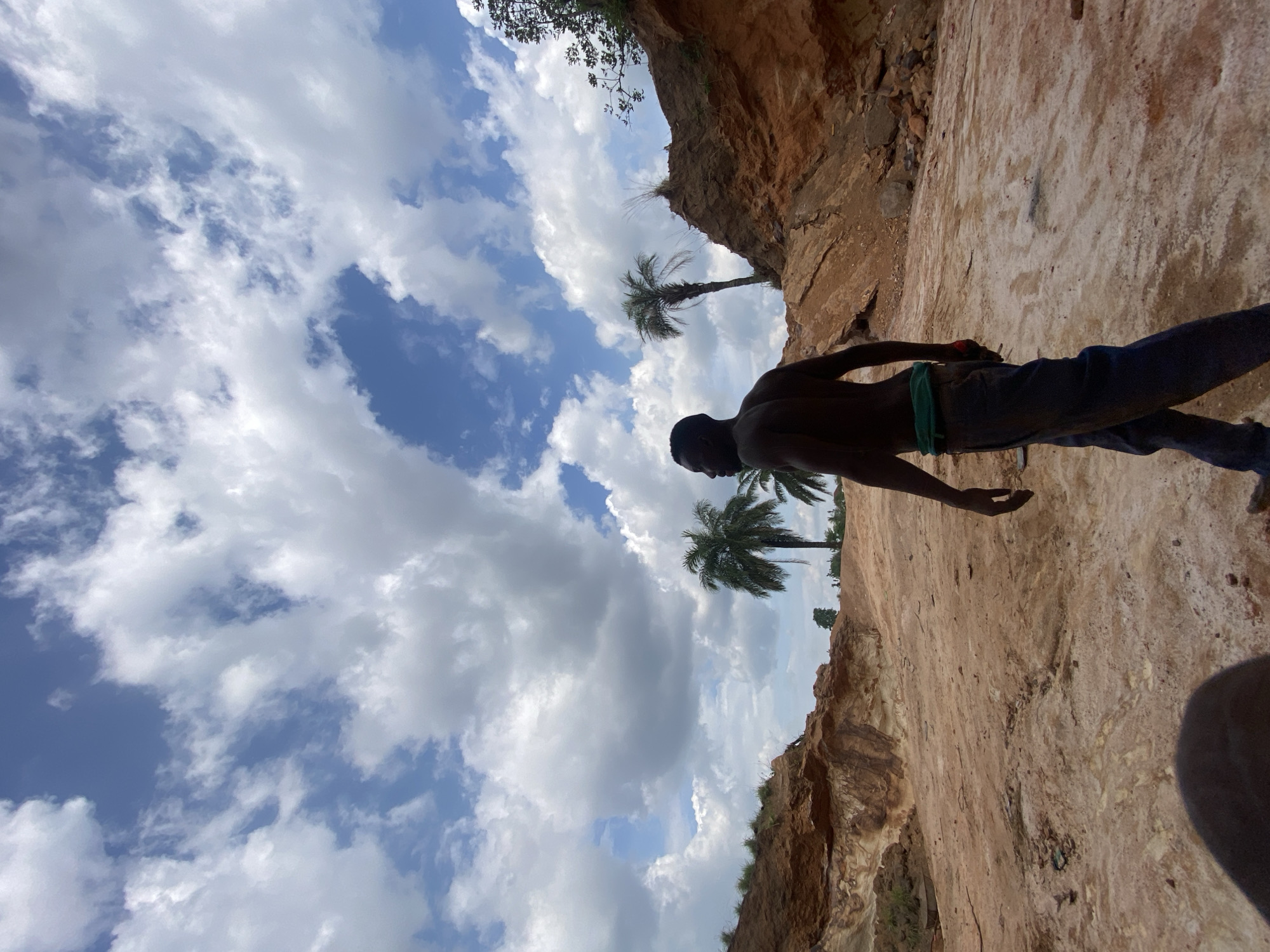 The scene depicts a shirtless Black man in jeans standing on a rocky, sandy terrain against a partly cloudy sky with palm trees. It appears to be outdoors, possibly in Igbara, Nigeria, given the context. The man is oriented as if walking or exploring the area, with the cliff face behind him. The sky occupies a significant portion of the frame, showcasing a mix of blue and white clouds.