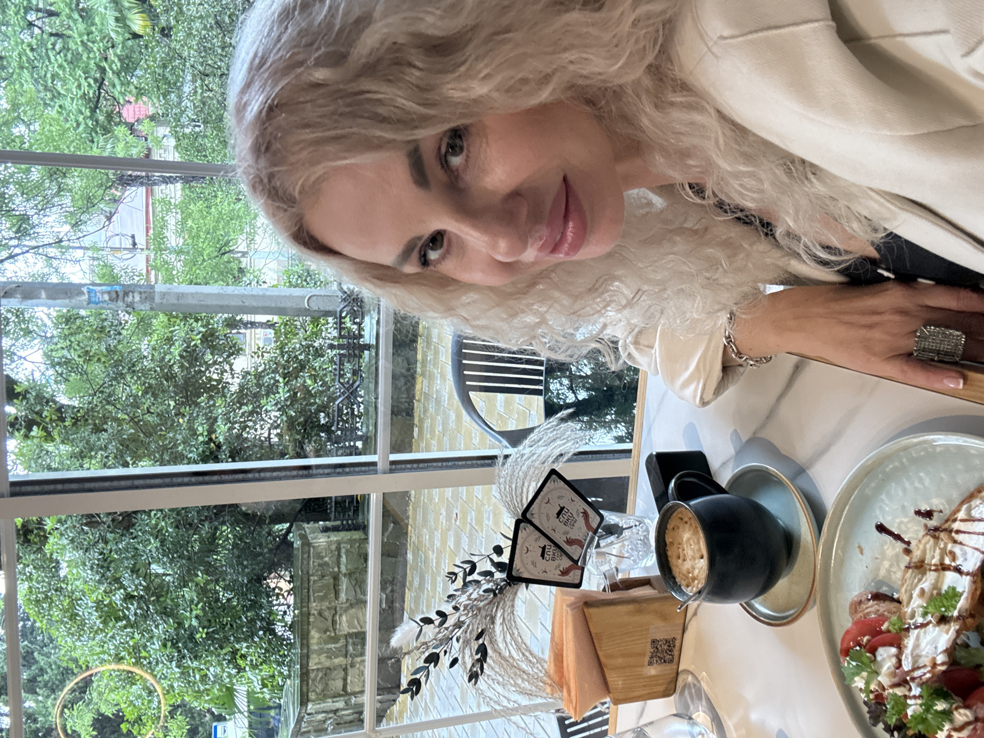 In Sochi, Russia, a blonde woman with curly hair is seated at a table inside a cafe, positioned to the right of the frame. She is wearing a white jacket and looking towards the camera with a slight smile. On the table in front of her is a cup of coffee with foam, a plate of food that appears to be a breakfast or brunch item with tomatoes and bread, and a wooden napkin holder. To the left of the coffee cup, there is a decorative arrangement of dried plants and two small cards with black and white graphics. Through the large window behind her, lush green trees and foliage are visible, suggesting an outdoor, natural setting surrounding the cafe. The weather appears overcast or rainy, as indicated by the diffused light.
