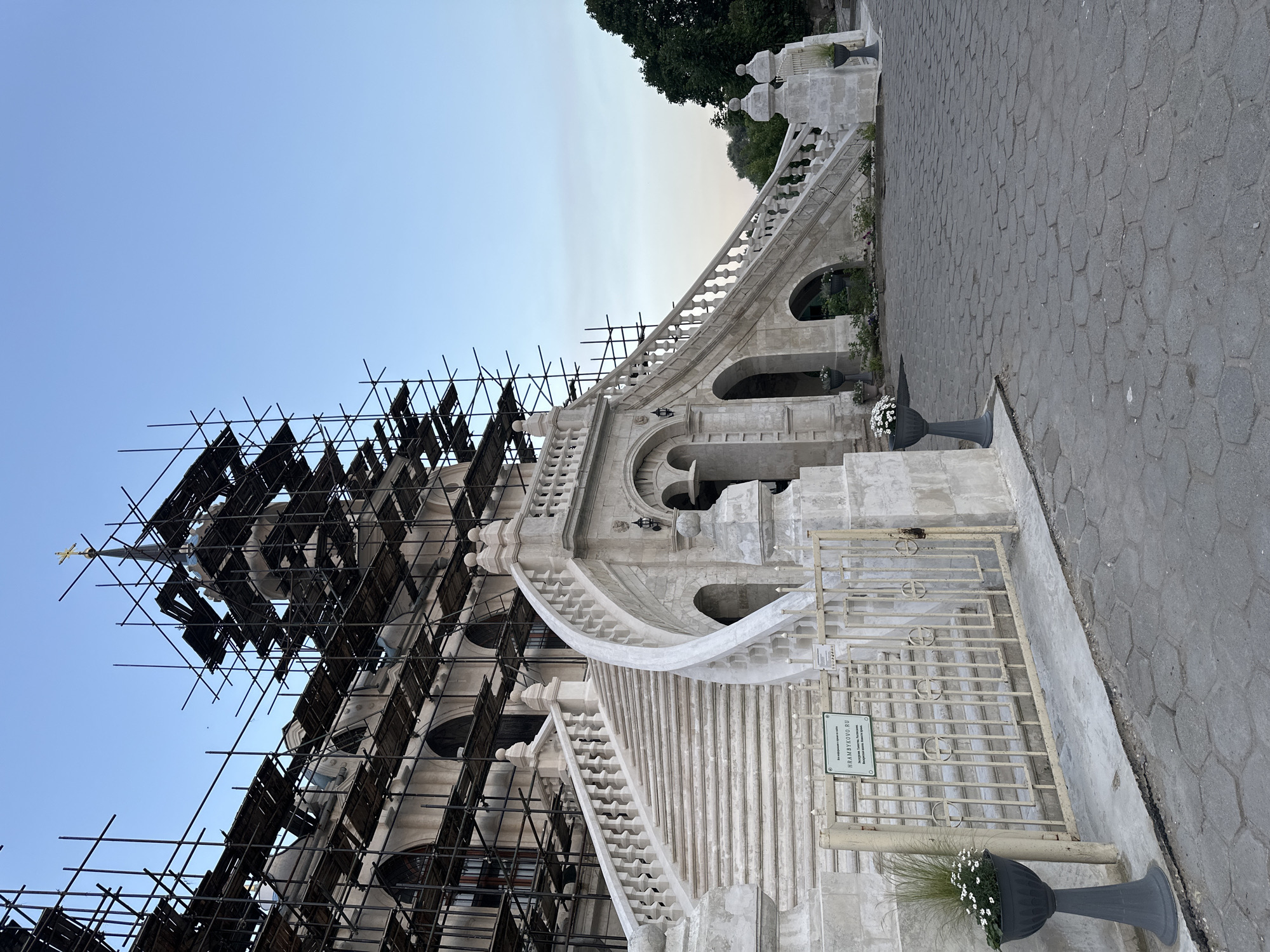 The image shows the Church of the Vladimir Icon of the Mother of God, located in the village of Bykovo, Russia, under renovation. A significant amount of scaffolding covers a large portion of the building's facade and spire. In the foreground, an elaborate white stone staircase with ornate balustrades curves upwards towards the main entrance. A metal gate with decorative circular motifs is situated at the bottom of the stairs, with a sign attached to it. Potted plants with white flowers are placed near the gate and along the walkway. The ground in front of the stairs is paved with grey hexagonal stones. The sky is a clear, pale blue, and the lighting suggests it is either early morning or late afternoon. No people are visible in the image.