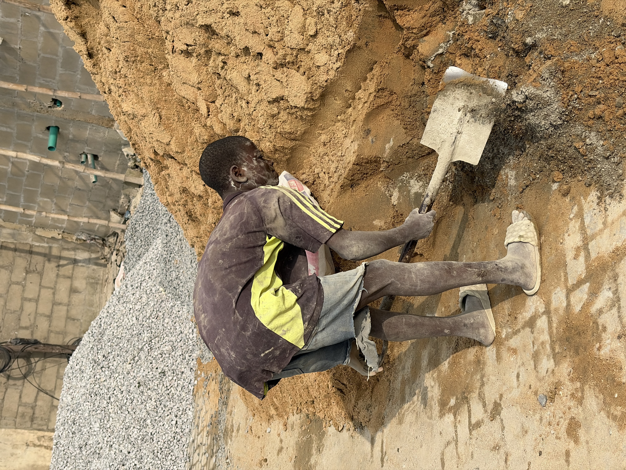 A person is shown working at a construction site in Igboefon, Nigeria. The individual is digging into a large pile of sand with a shovel. They are wearing a dirty, dark t-shirt with a yellow stripe, denim shorts, and sandals. The environment is dusty with piles of sand and gravel visible. In the background, a brick wall with exposed pipes and bamboo scaffolding suggests ongoing construction work. The lighting indicates it is daytime.