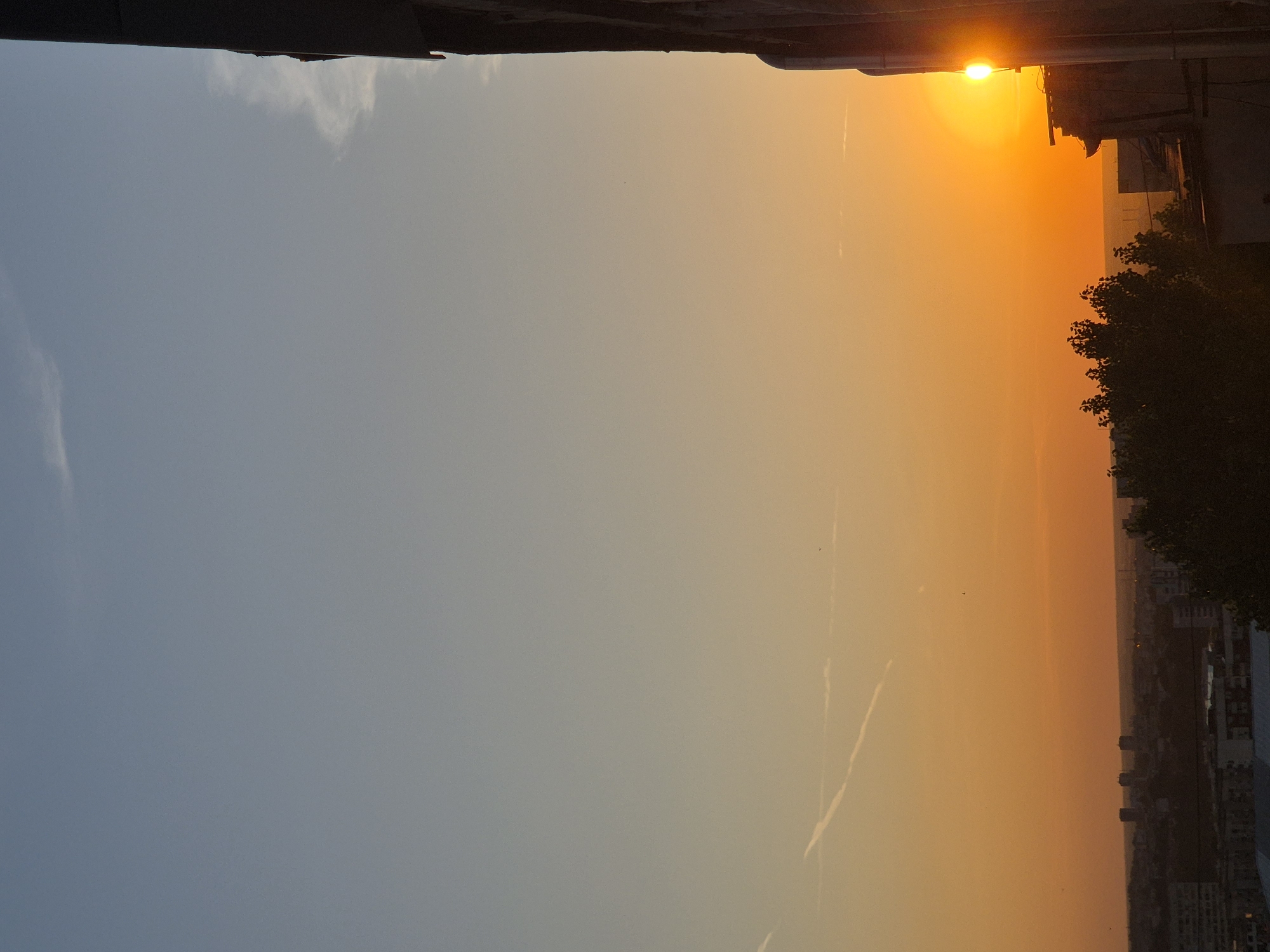 The image captures a city skyline at sunset. The sky transitions from a pale blue at the top to a vibrant orange as it nears the horizon. A faint wispy cloud stretches across the upper portion of the sky, and a contrail from an aircraft is visible lower down. The sun, a bright golden orb, is partially obscured by a building on the right side of the frame, casting a warm glow. Silhouetted against the sunset is a dense cluster of trees in the mid-ground. Beyond the trees, a cityscape with various buildings can be seen stretching to the horizon. The foreground is dominated by the side of a building, also in silhouette, with vertical elements that appear to be pipes or structural supports. The overall scene depicts a serene urban sunset.