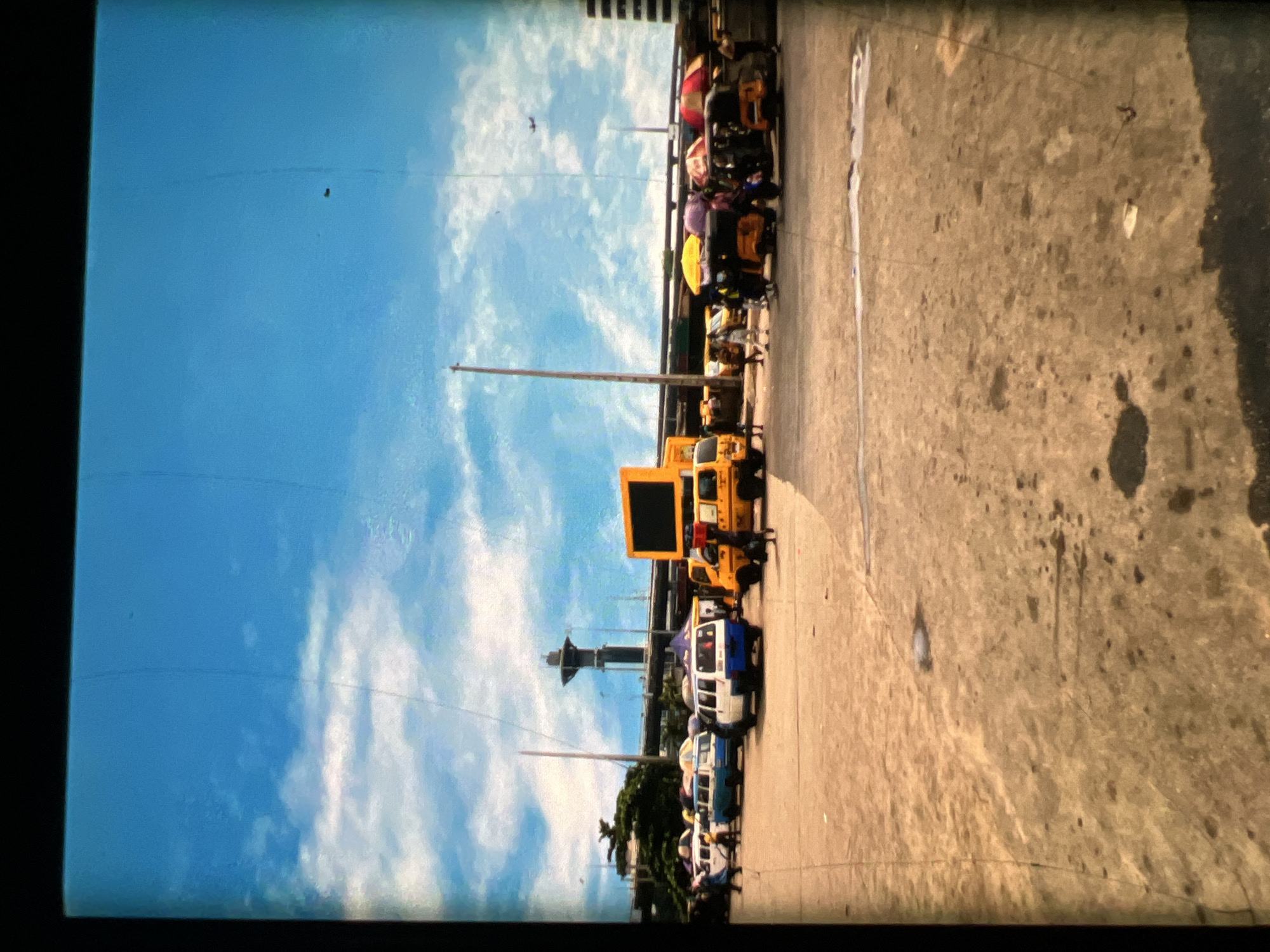 This image depicts a bustling street scene in Lagos, Nigeria, likely a transportation hub or market area. The foreground is dominated by a paved, somewhat weathered surface, showing signs of wear and some damp patches. In the mid-ground, various vehicles are parked or moving. Several white minibusses, common as public transport in Lagos, are visible. There are also yellow open-sided vehicles, possibly commercial vans or specialized transport. Several colorful umbrellas, red and yellow, are set up over some of the vehicles, offering shade. A large yellow billboard with a black screen is prominently displayed, likely used for advertising. People are scattered throughout the scene, some standing near the vehicles, others walking. In the background, a distinctive tower structure with a dark cap is visible, possibly a landmark or part of a larger infrastructure. Overhead, a bright blue sky with scattered white clouds suggests fair weather. A faint airplane can be seen in the sky. The overall impression is one of a busy, active urban environment characteristic of Lagos.